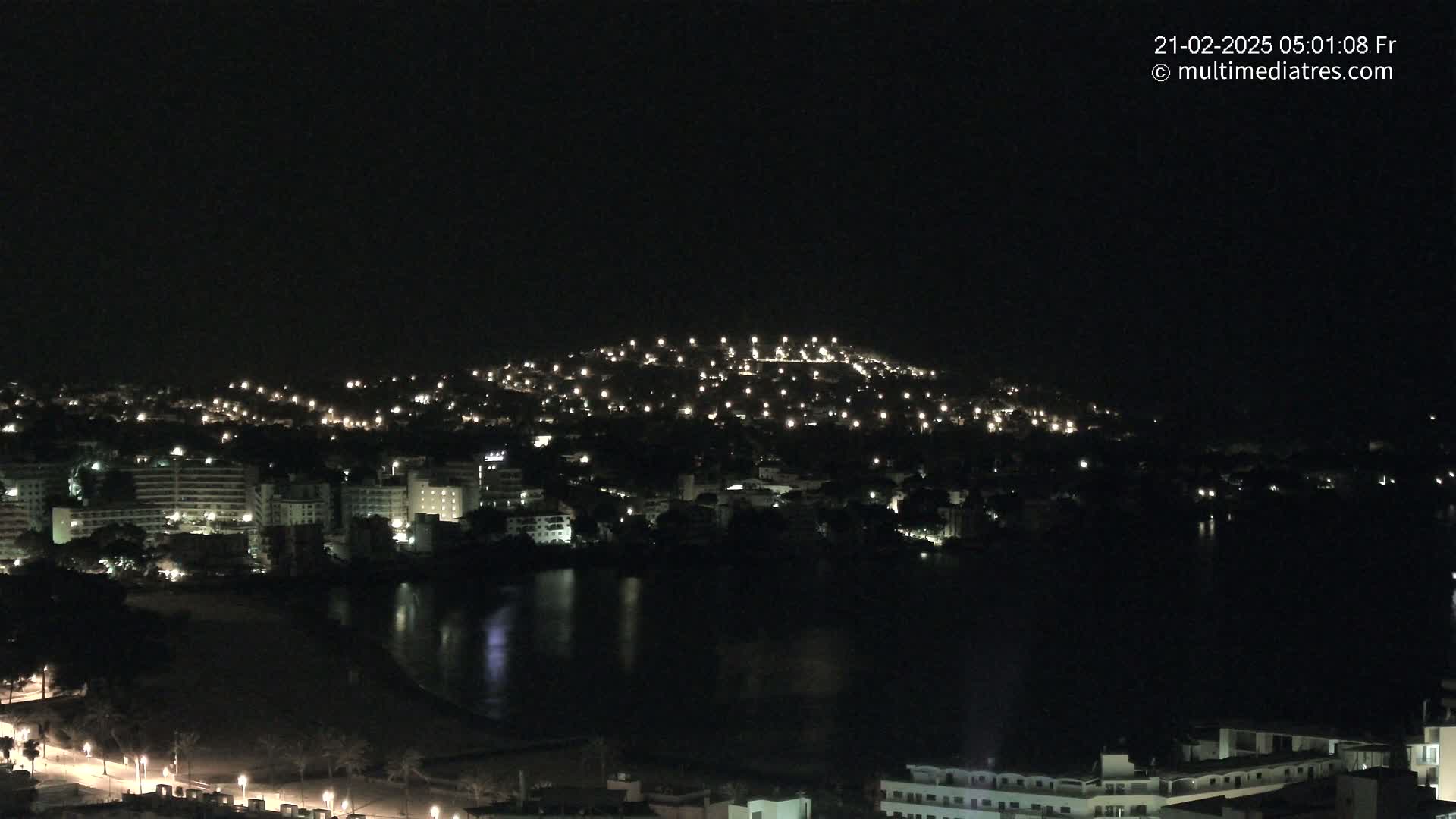 A nighttime view of a city sprawling across a hill and alongside a dark body of water, illuminated by scattered lights.
