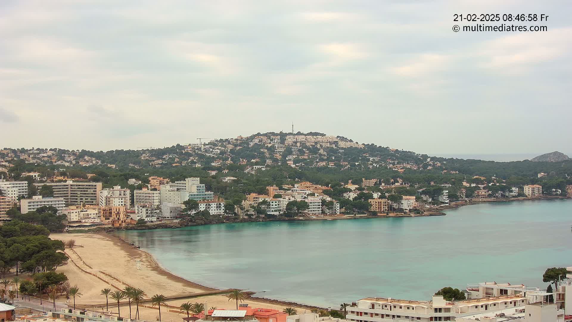 A coastal town with buildings nestled on a hillside overlooking a calm, turquoise bay with a sandy beach under a cloudy sky.