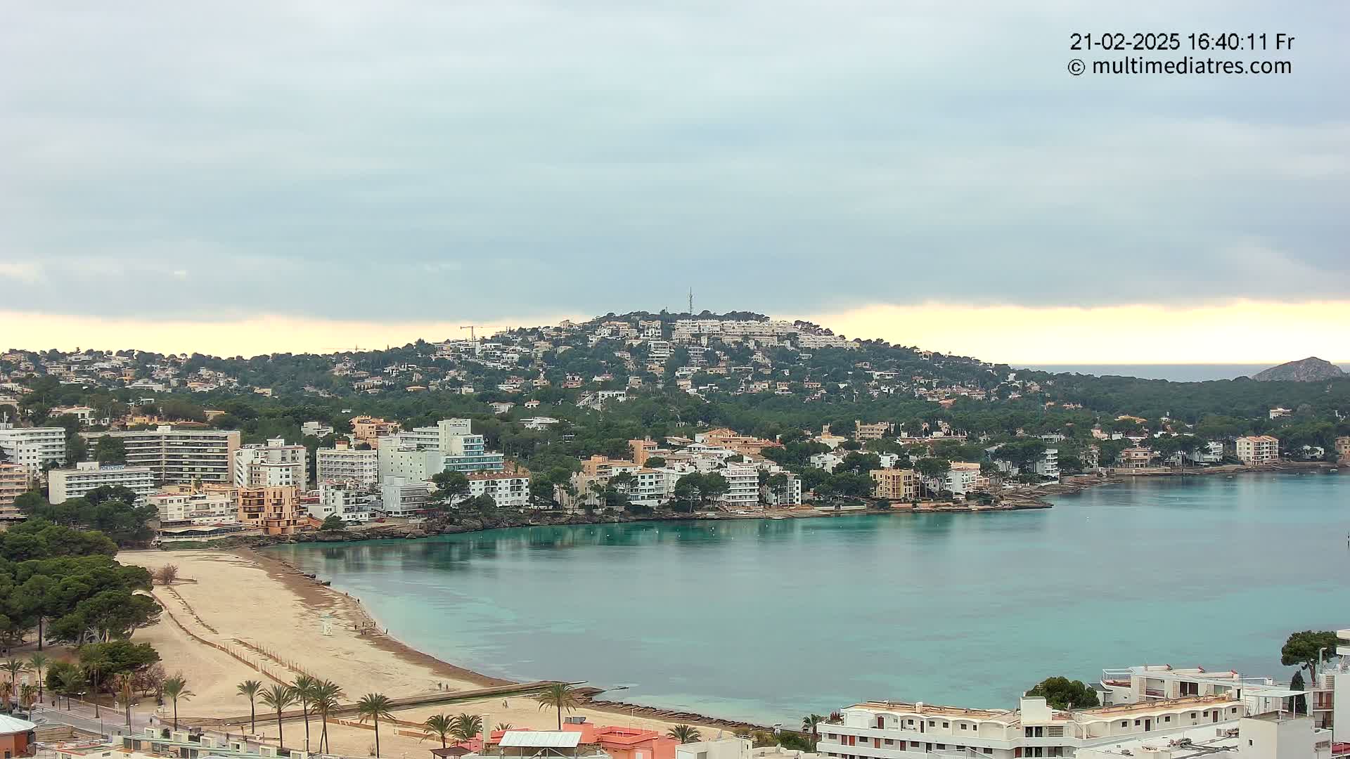 A coastal town with numerous buildings sprawls across a hillside and along a bay with a sandy beach, under an overcast sky.
