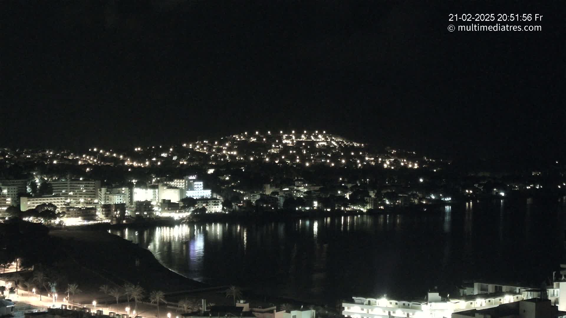 A nighttime view of a coastal town illuminated with many lights, reflected in a calm body of water.