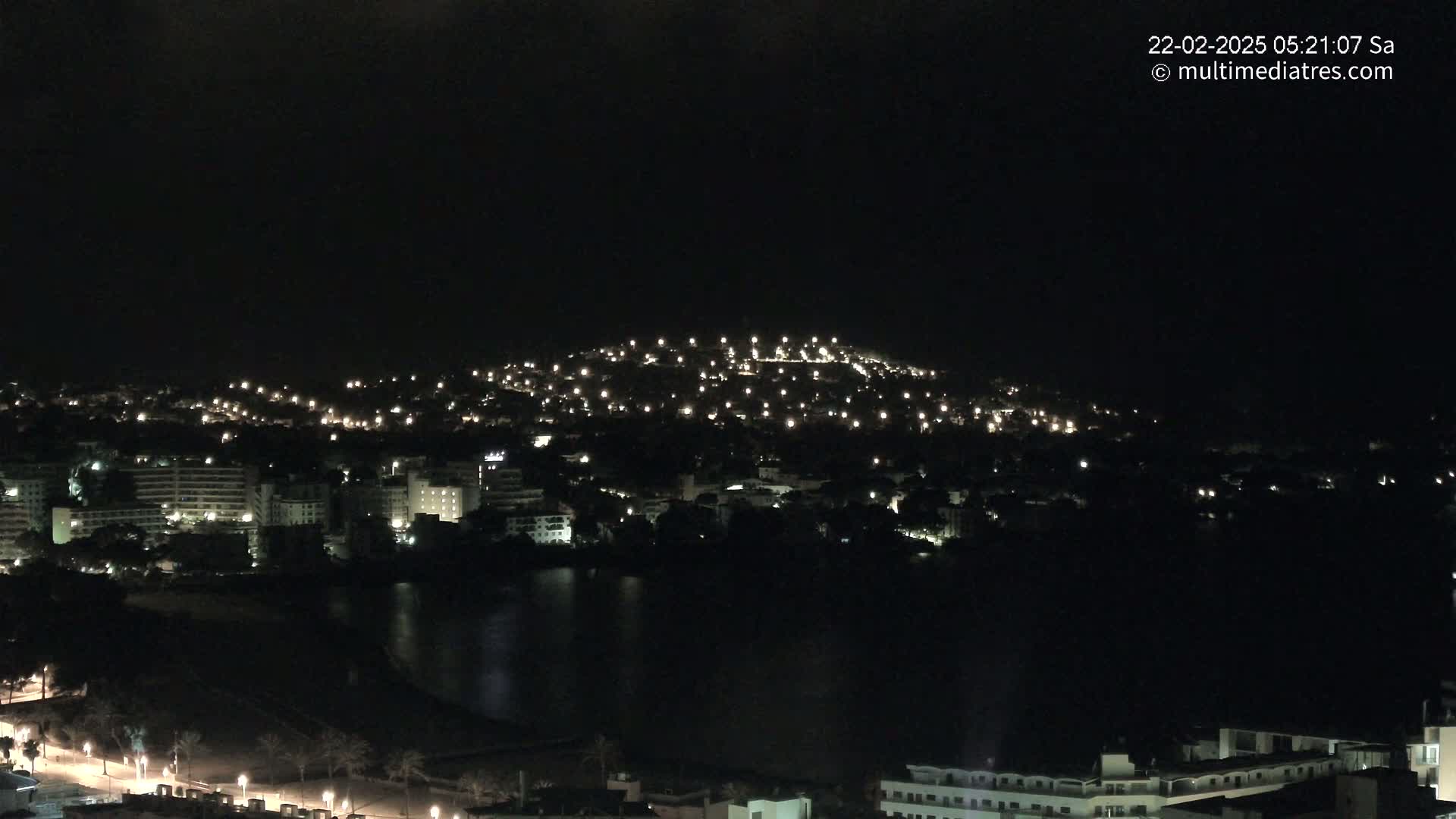 A nighttime view of a city nestled around a body of water, illuminated by numerous lights on buildings and hills.