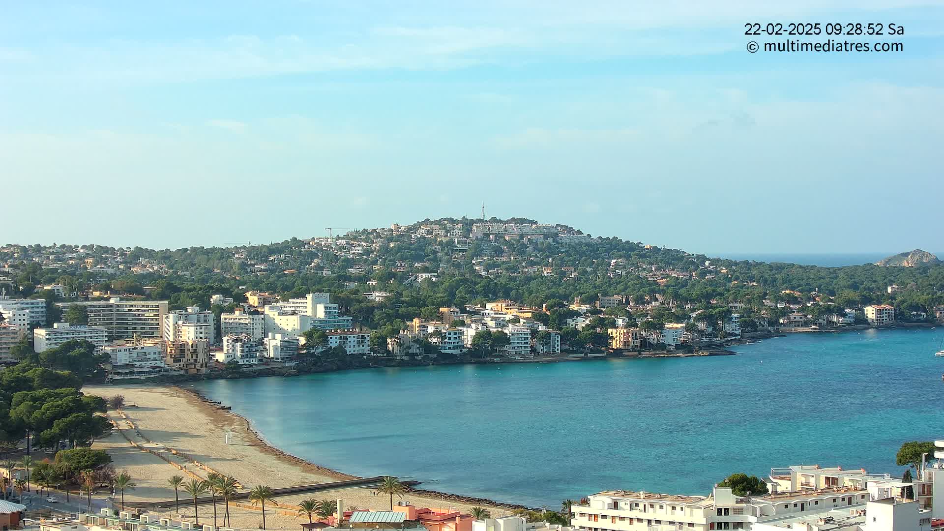 A coastal town with a sandy beach and turquoise water under a mostly clear sky nestles against a hill covered in buildings and trees.