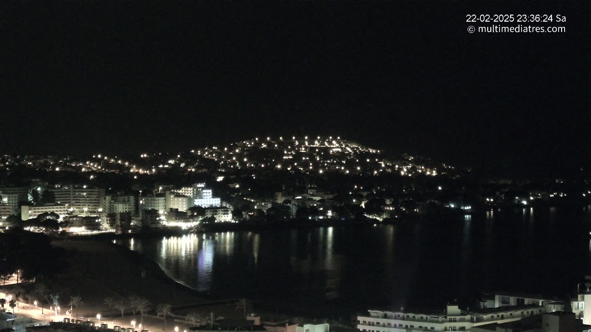 A nighttime view of a coastal town with numerous lights illuminating buildings and streets, reflected in a calm body of water.