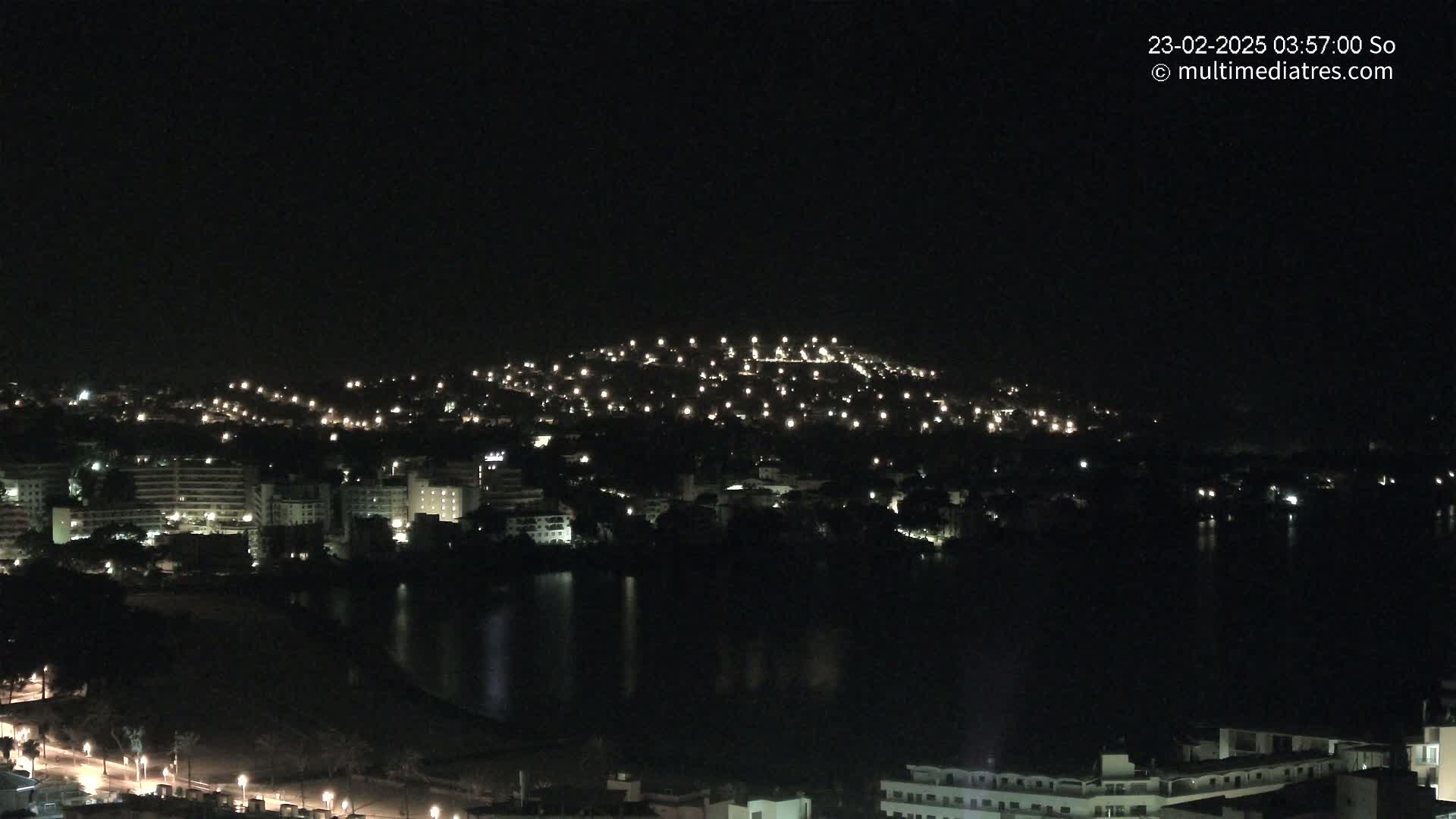 A nighttime view of a city illuminated by numerous lights, situated beside a dark body of water, with a hilltop also speckled with lights in the background.