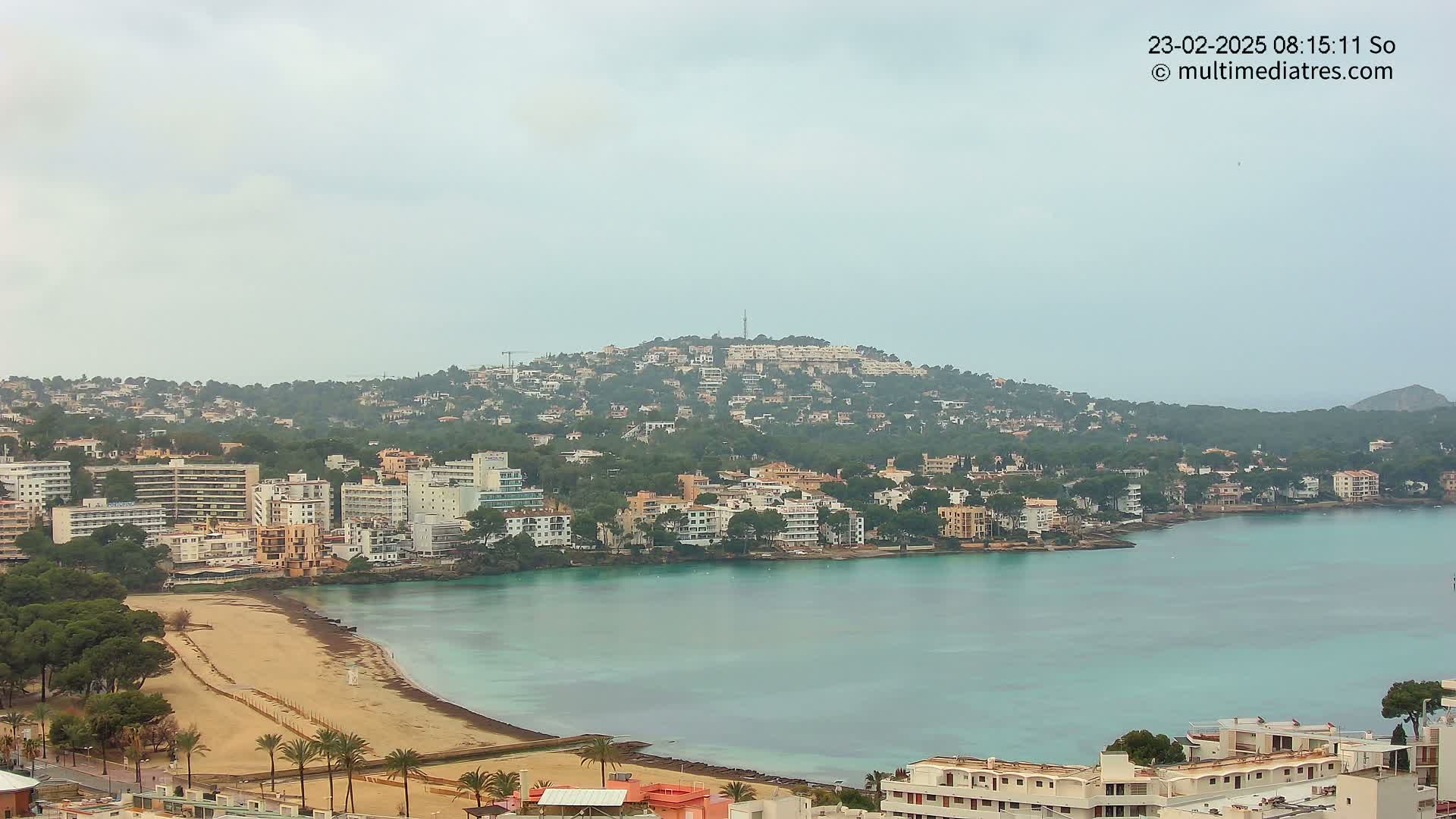 A coastal town with a sandy beach and numerous buildings stretches across a hillside overlooking a calm, teal bay under an overcast sky.