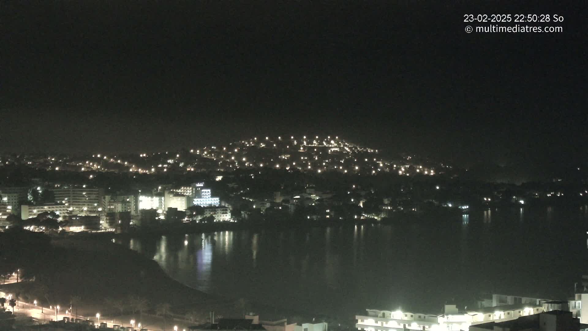 A nighttime view of a coastal town with numerous lights illuminating buildings and a hill overlooking a calm body of water.