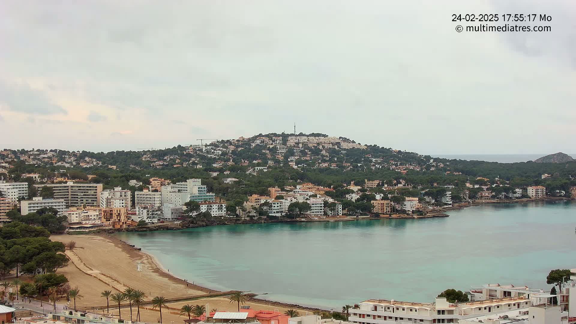 A coastal town nestled on a hillside, with a sandy beach and calm turquoise waters under an overcast sky.