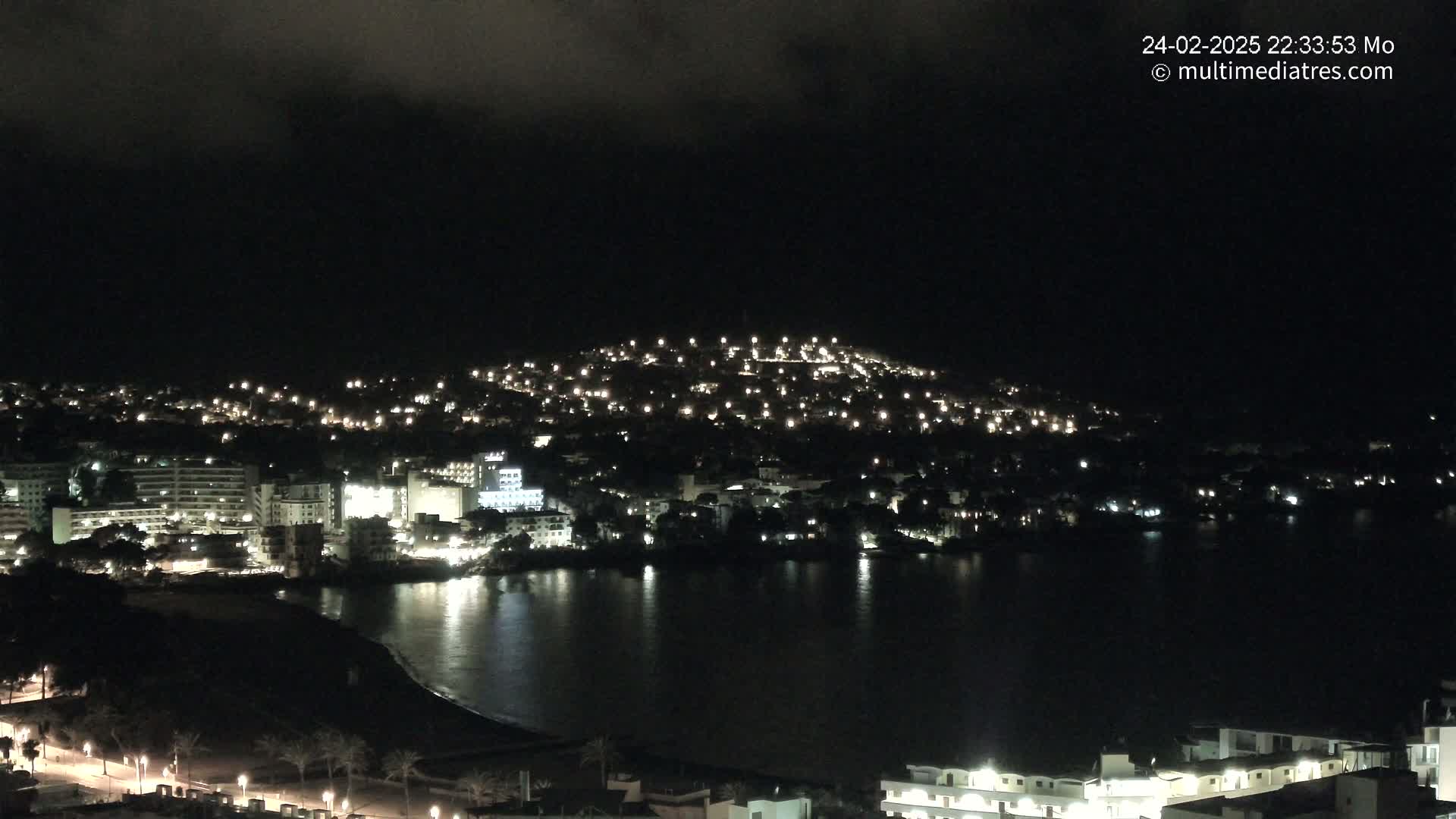 A nighttime view of a coastal town illuminated by lights, under a dark, cloudy sky.