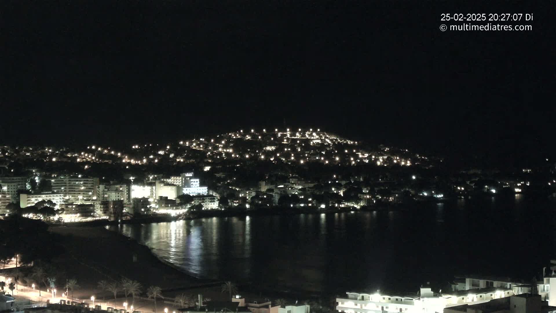 A nighttime view of a coastal town illuminated by many lights, with calm dark water reflecting the city lights.