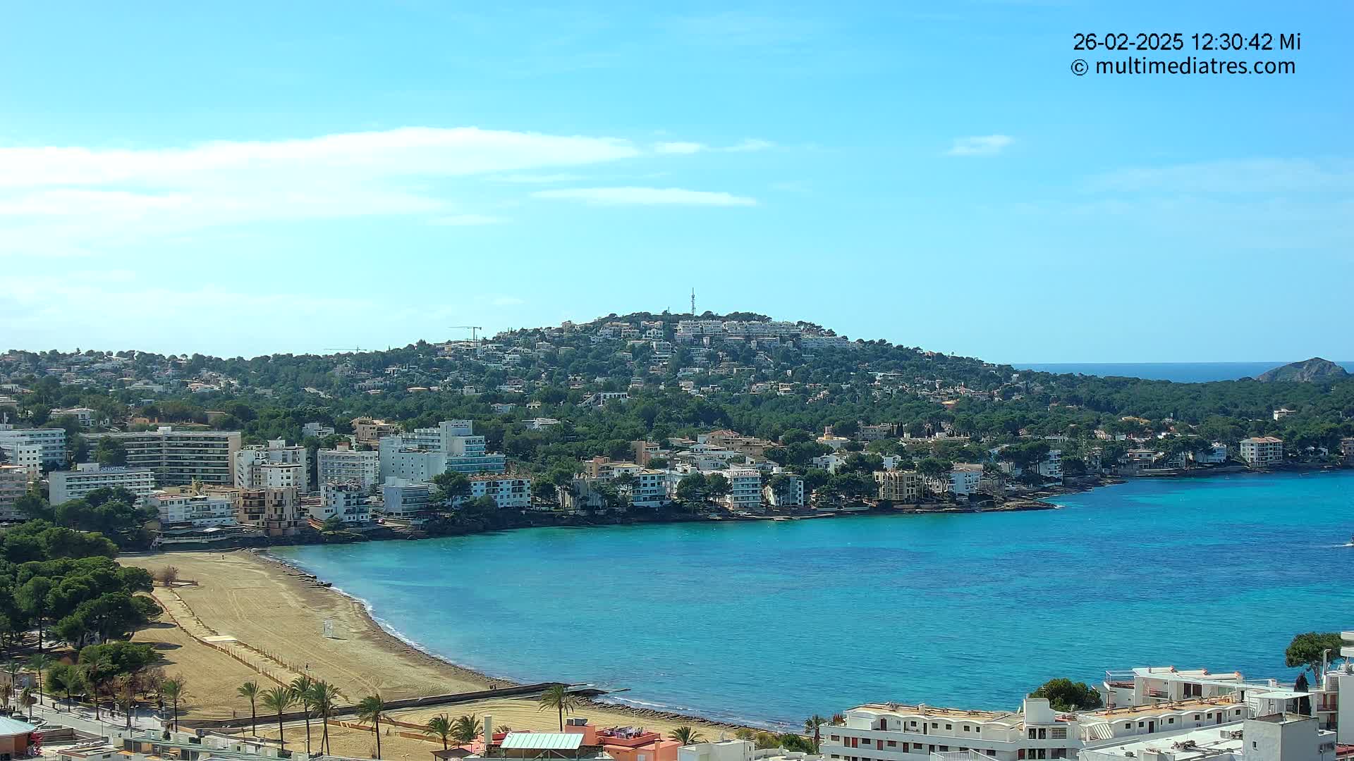 A coastal town with sandy beach, turquoise water, and buildings climbing a verdant hill under a clear blue sky.