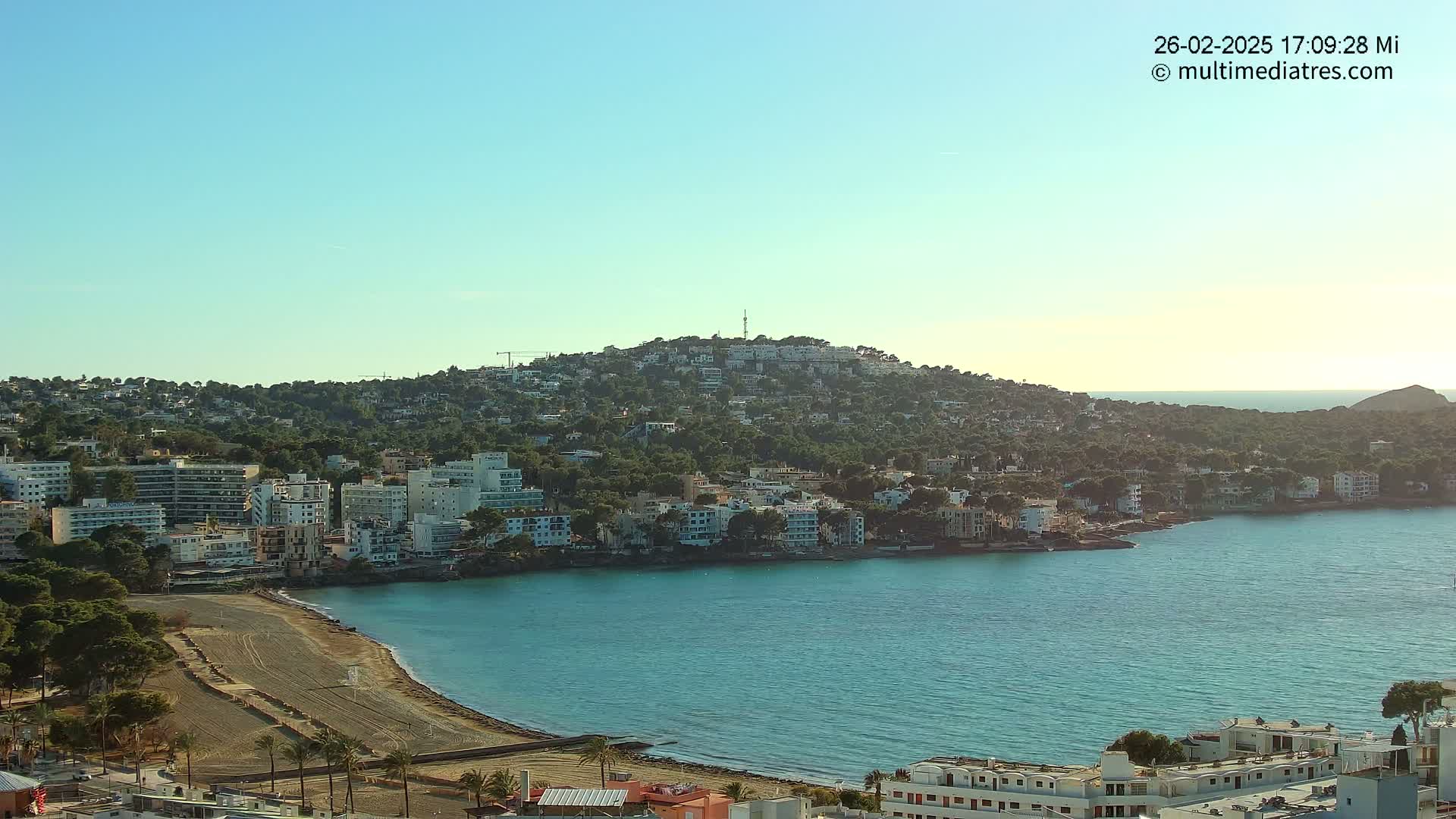 A coastal town nestled on a hillside overlooking a calm, turquoise bay under a clear, bright sky.