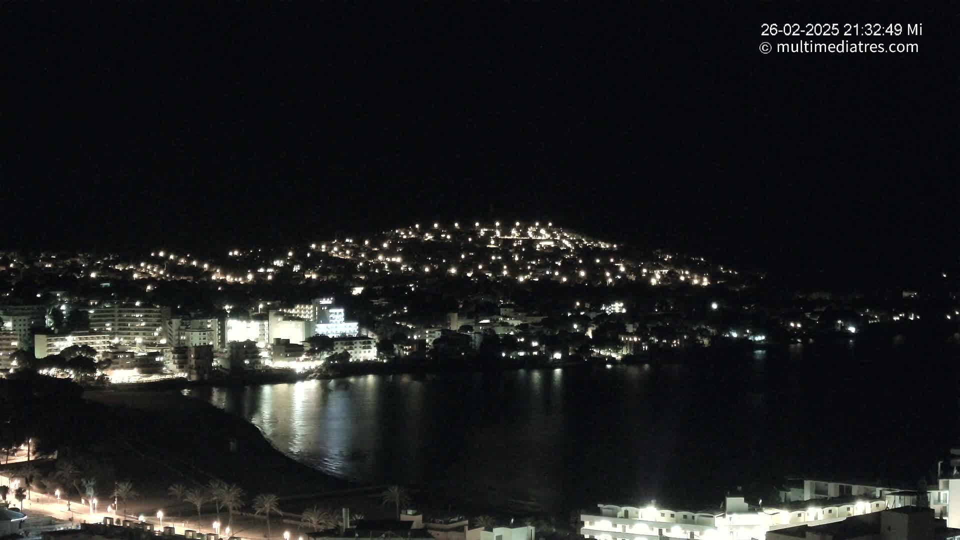 A nighttime view of a coastal town illuminated by many lights, situated on a hill and beside a dark body of water.