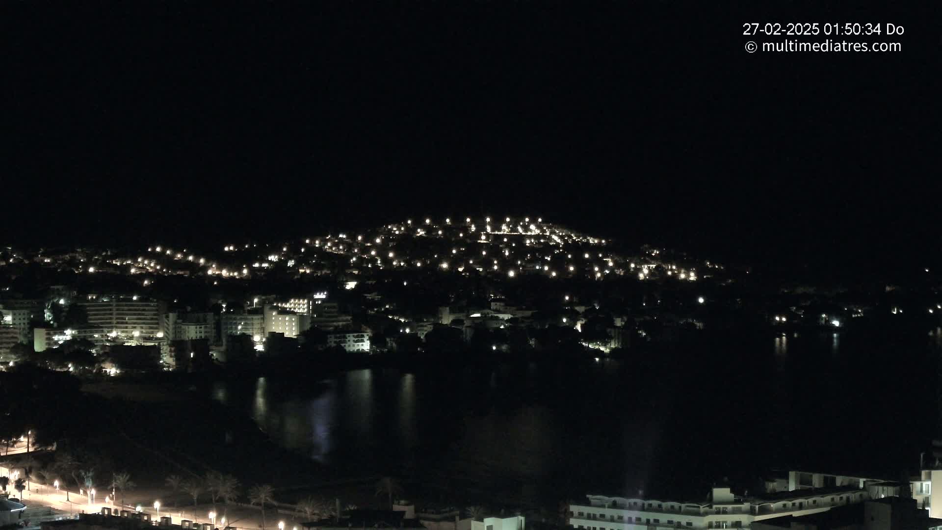 A nighttime view of a coastal town illuminated by many lights, with a dark body of water reflecting some of the light.