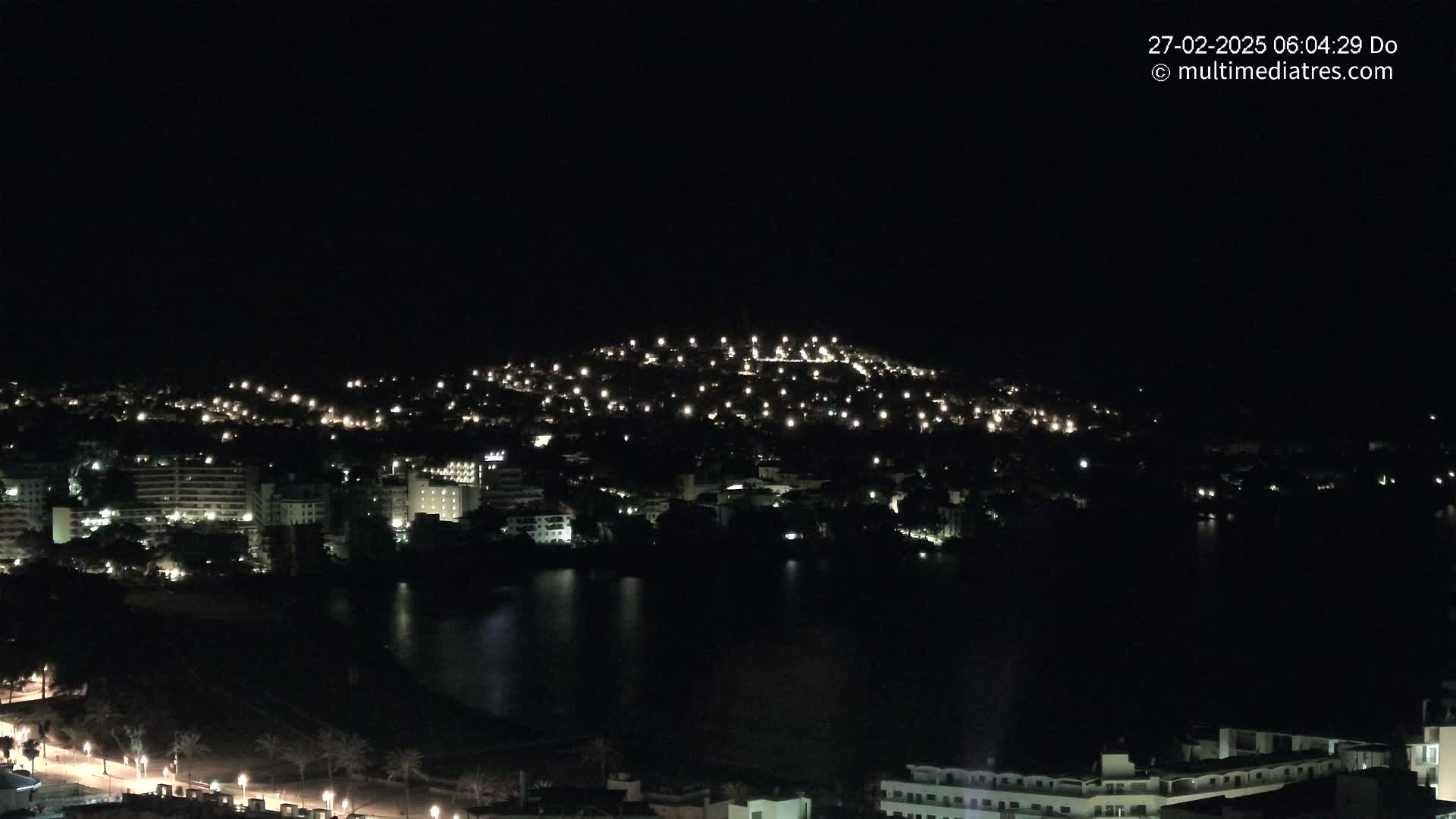 A nighttime view of a city illuminated by lights, nestled beside a dark body of water, under a clear, dark sky.