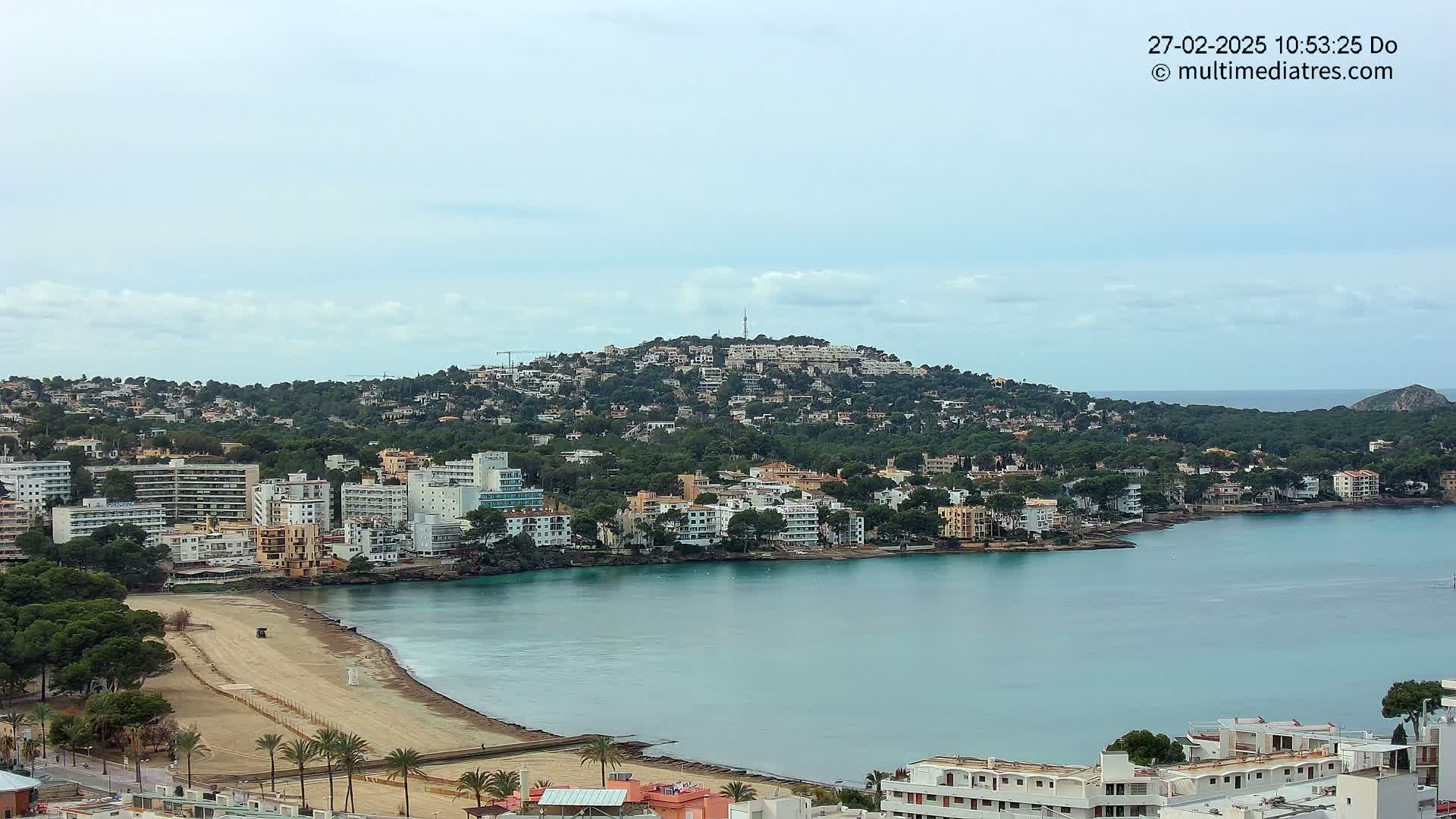 A coastal town with buildings nestled along a hillside and a sandy beach is seen under a mostly cloudy sky.