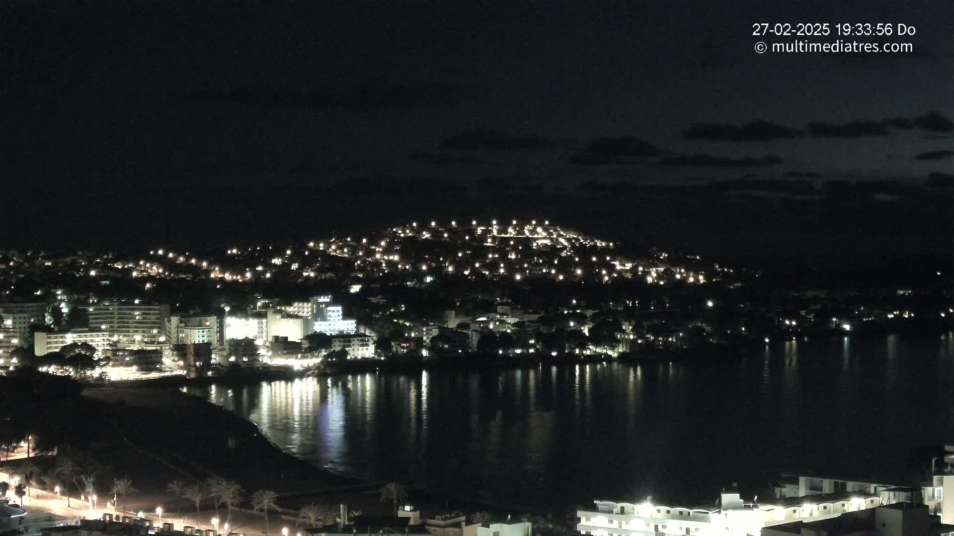 A nighttime view of a coastal town illuminated by many lights, reflected in a calm body of water under a dark sky.
