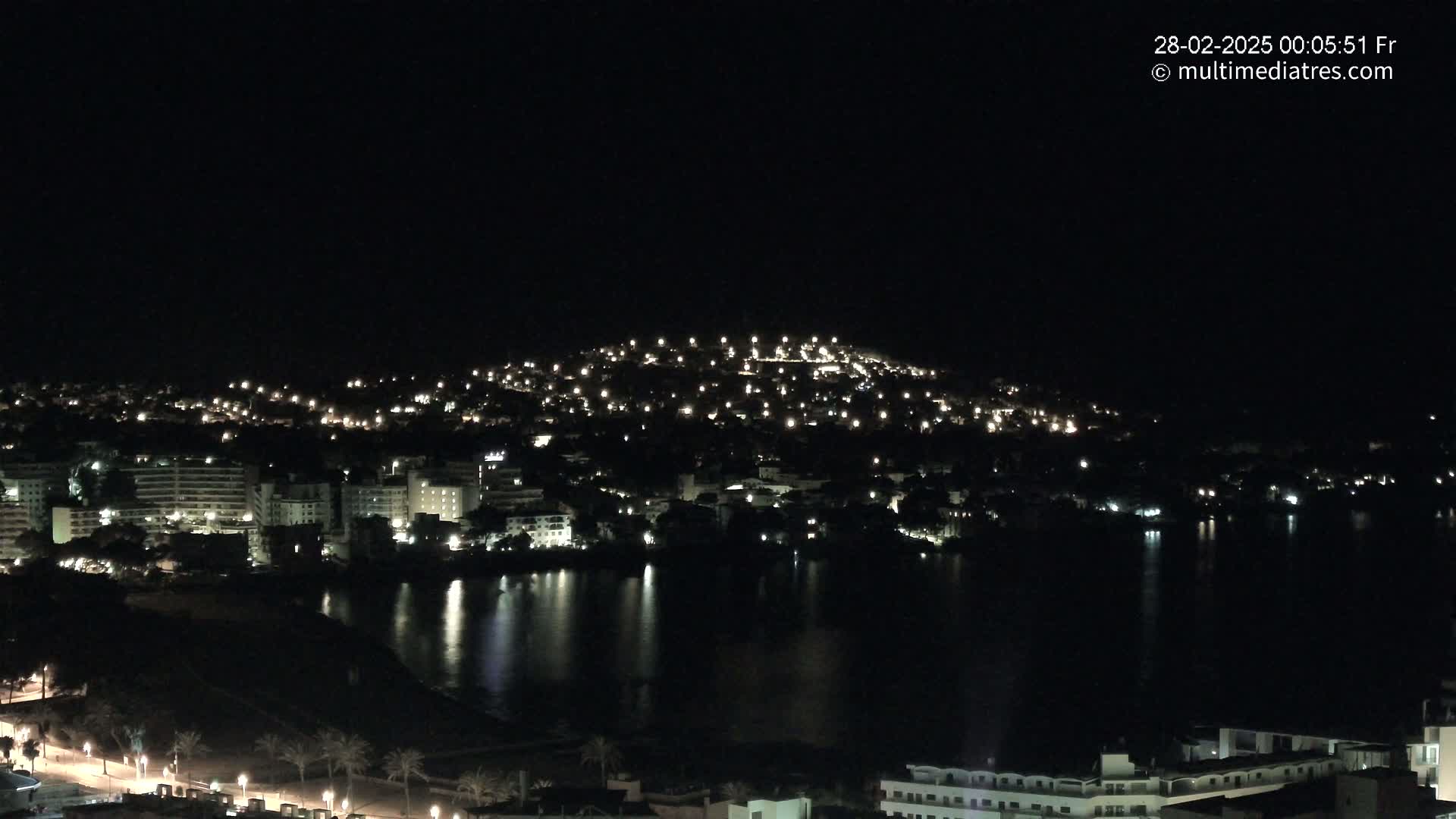 A nighttime view of a coastal town with buildings illuminated along a dark body of water under a clear, dark sky.