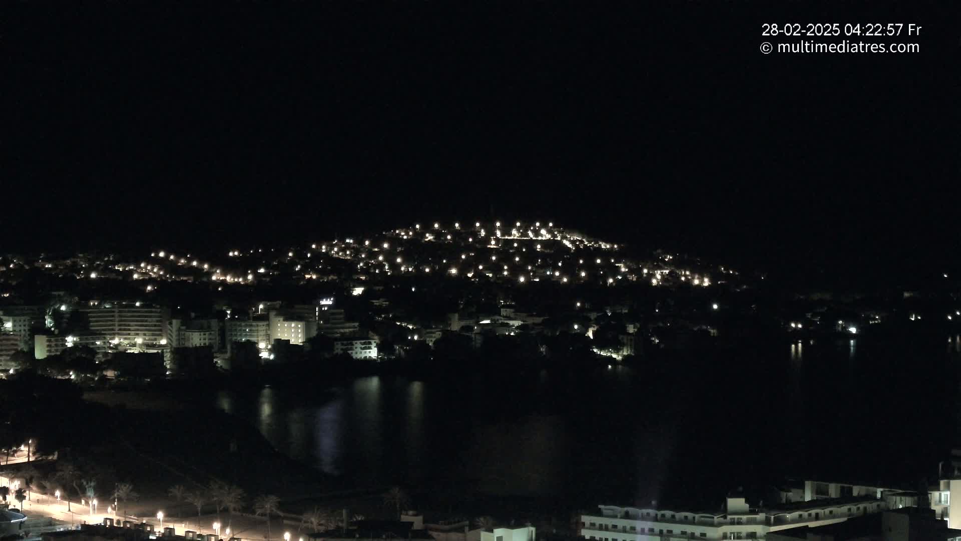 A nighttime view of a city nestled beside a dark body of water, illuminated by scattered lights.