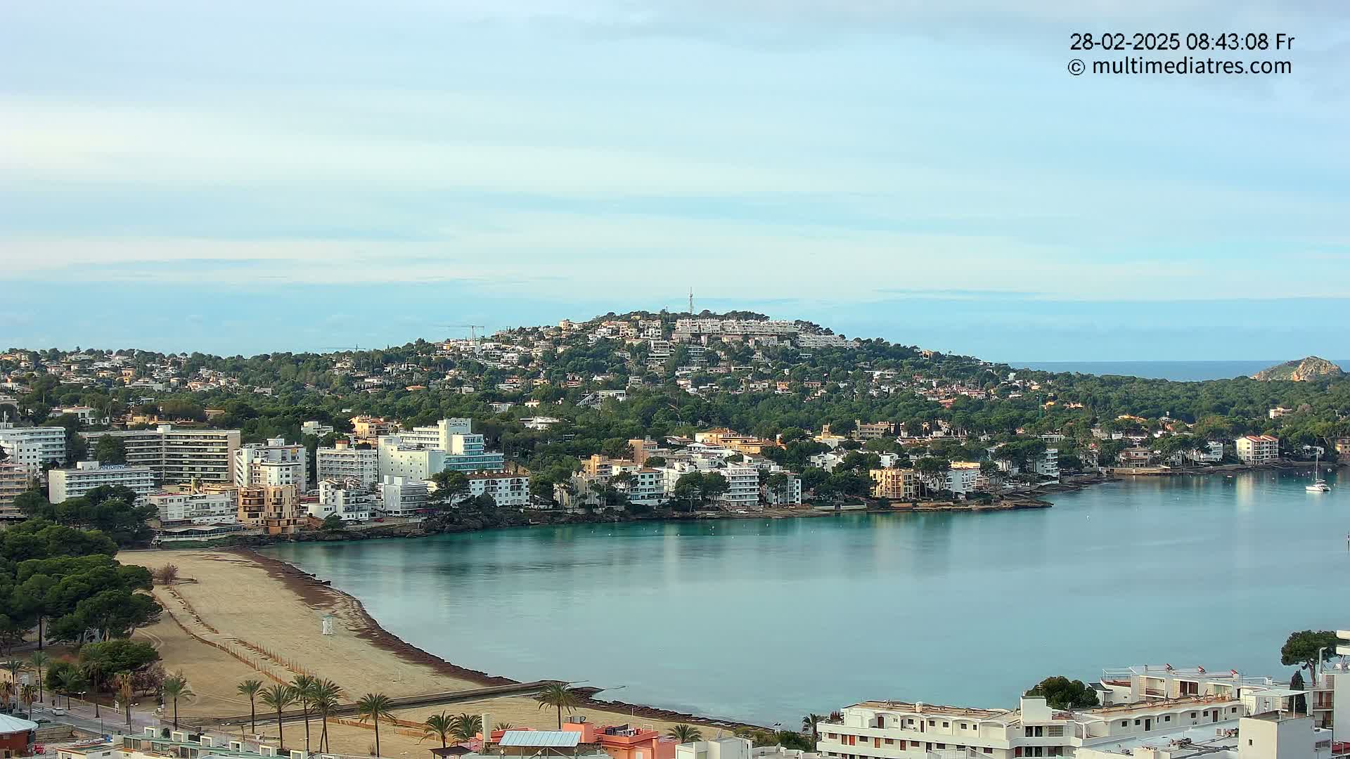 A coastal town with buildings clustered around a calm, teal bay, under a mostly clear sky.