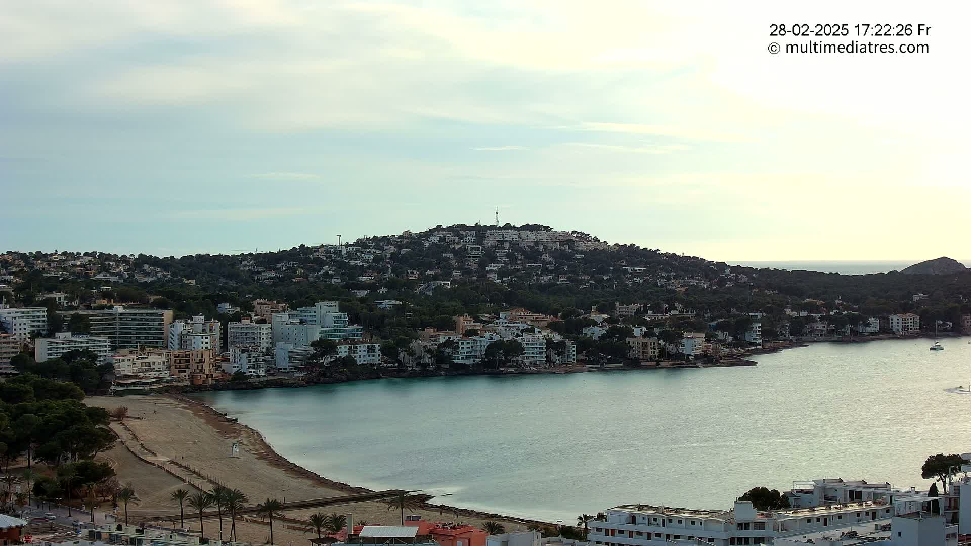 A coastal town nestled against a hill, with a calm bay in front, under a cloudy sky.