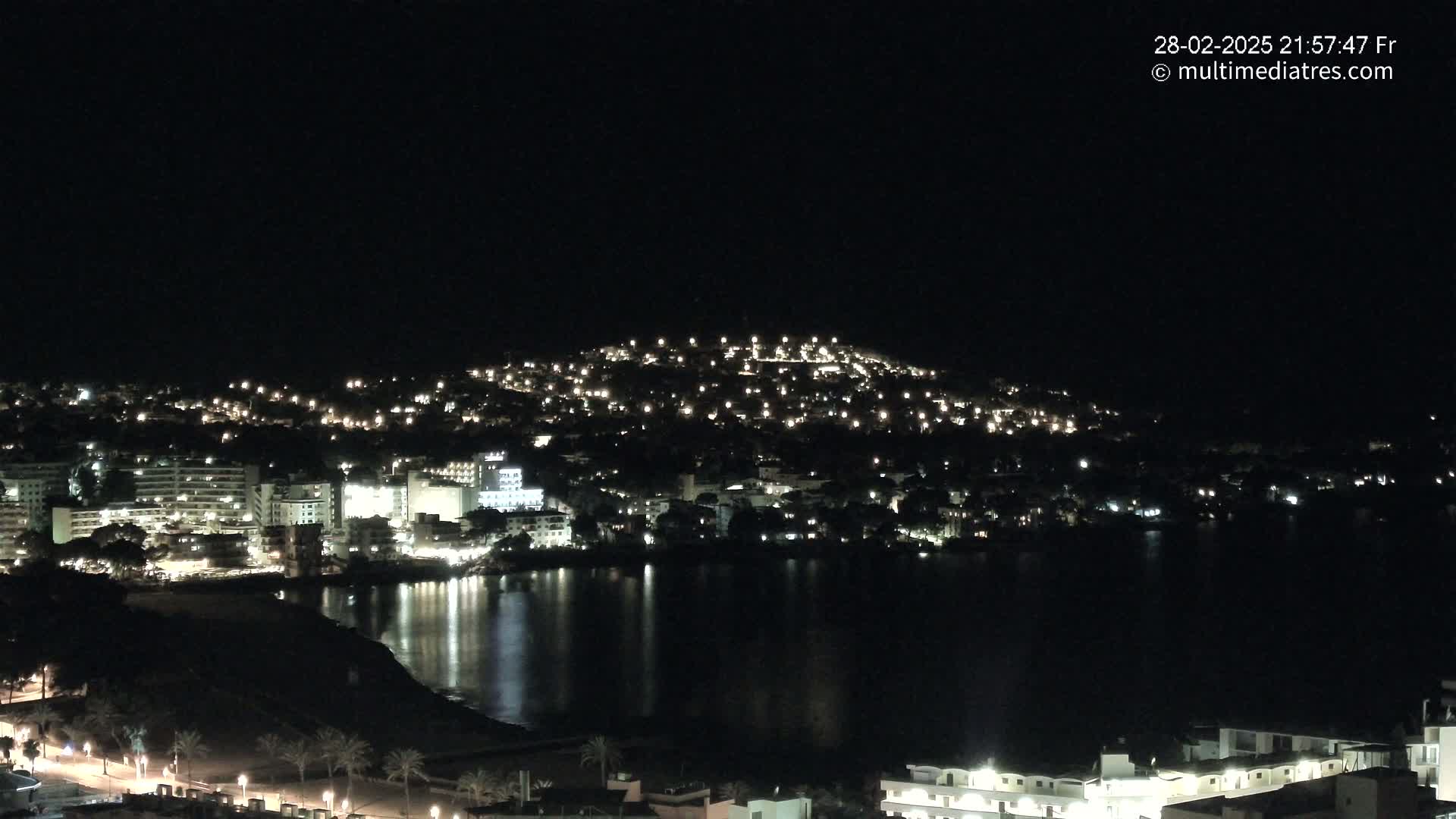 A nighttime view of a coastal town illuminated by many lights, with a dark, calm body of water reflecting the city lights.