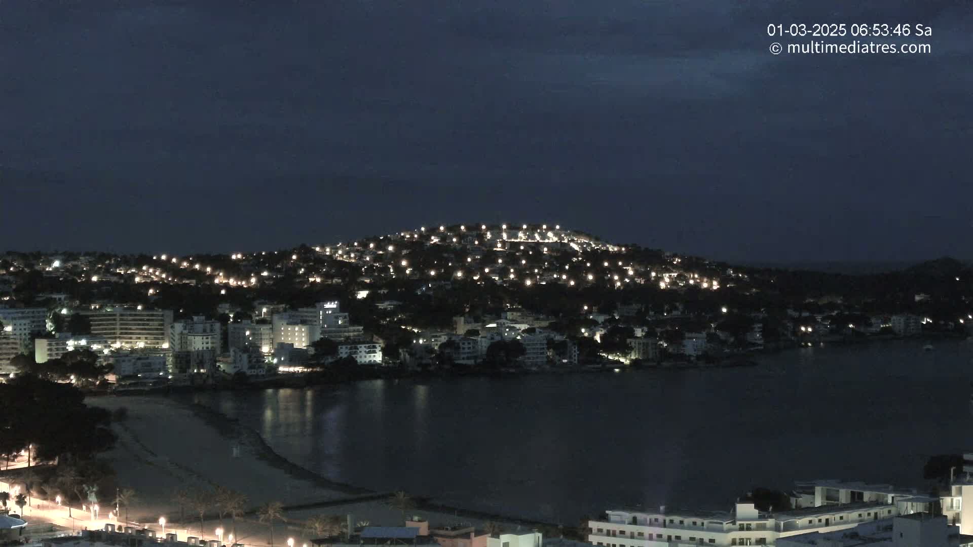 A nighttime view of a coastal town illuminated by many lights, situated on a hill overlooking a dark body of water and a sandy beach under a dark, clear sky.