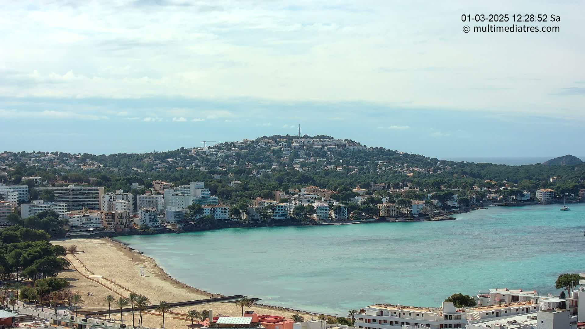 A coastal town nestled against a verdant hill, with a sandy beach curving into a turquoise bay under a partly cloudy sky.