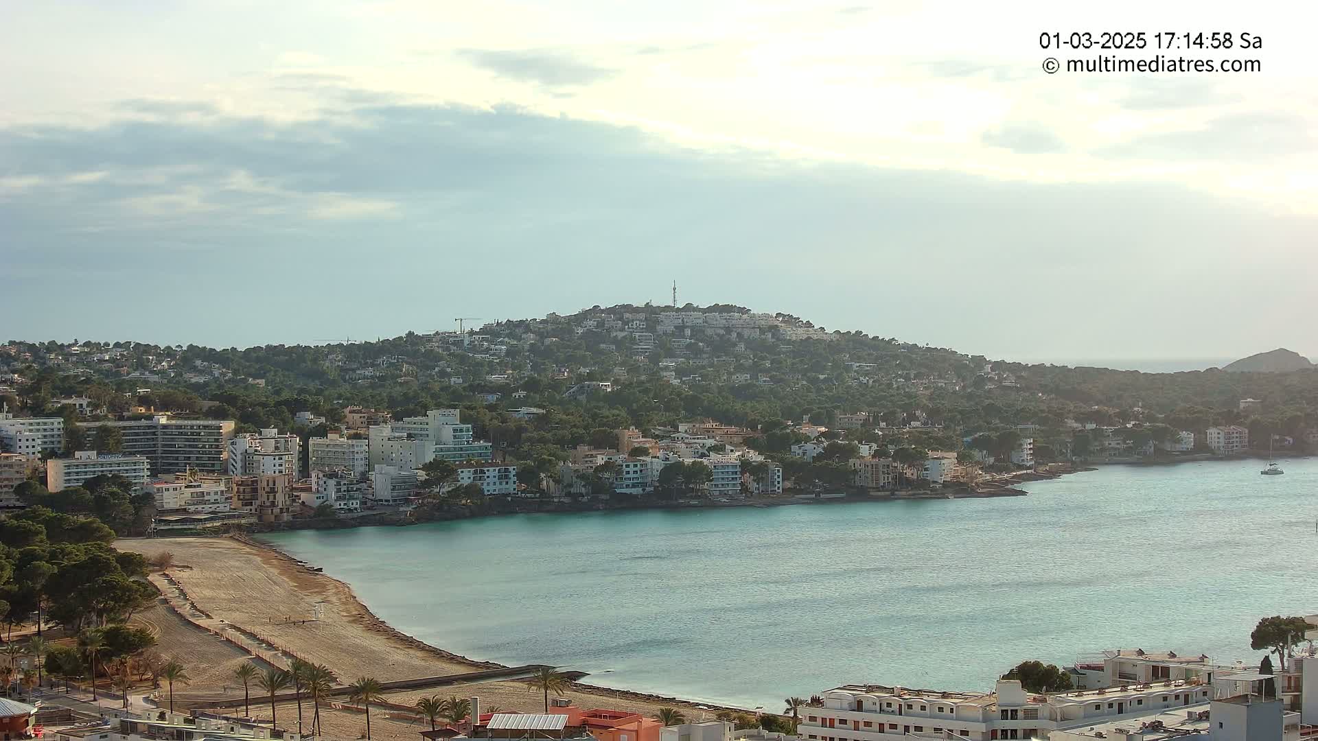 A coastal town nestled below a hill dotted with buildings, under a partly cloudy sky, features a calm, turquoise bay with a sandy beach area.