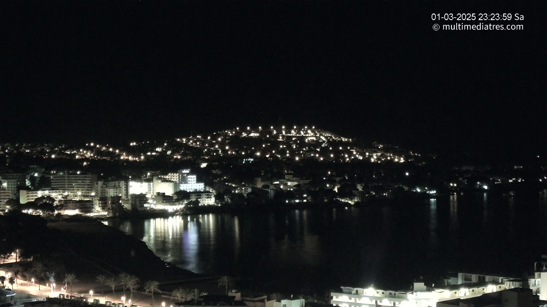 A nighttime view of a coastal town with its lights reflecting on a calm body of water.