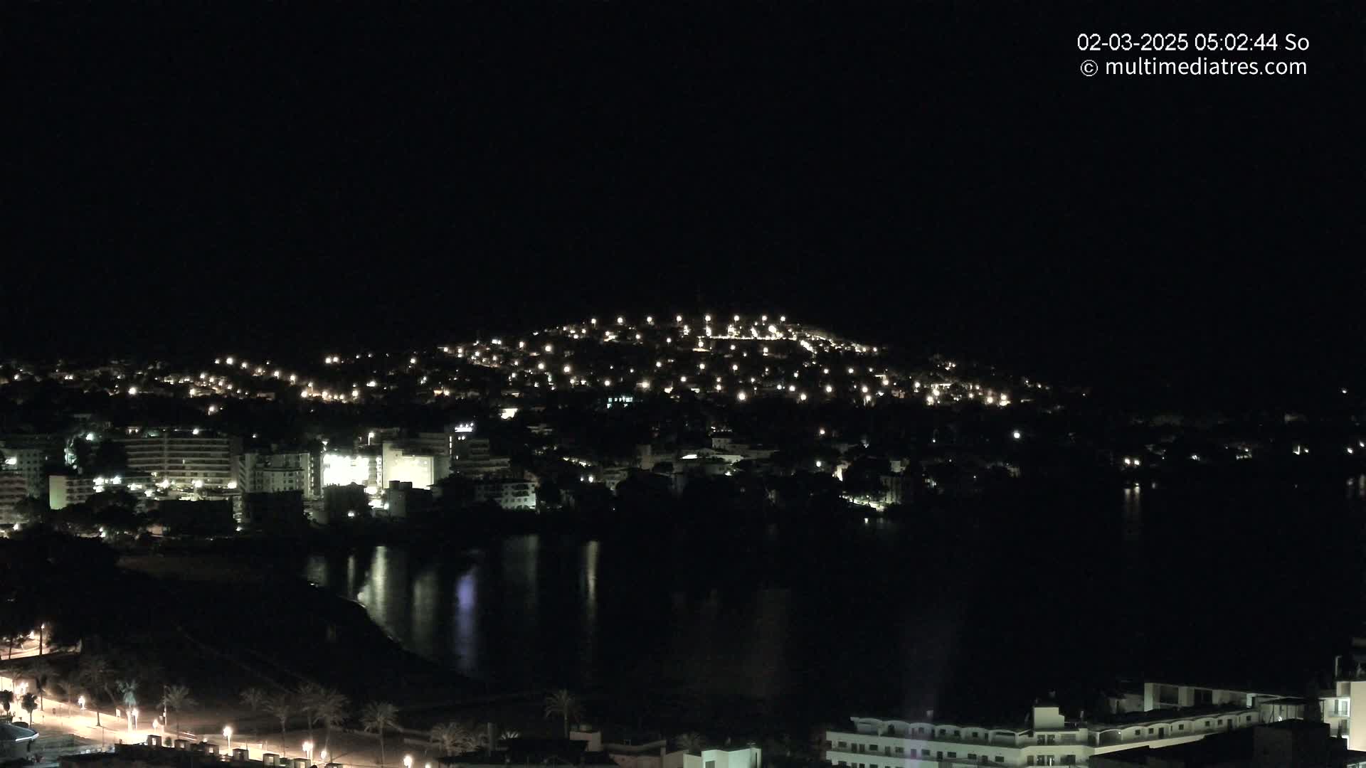 A nighttime view of a coastal town with buildings lit up along a dark body of water under a clear, dark sky.