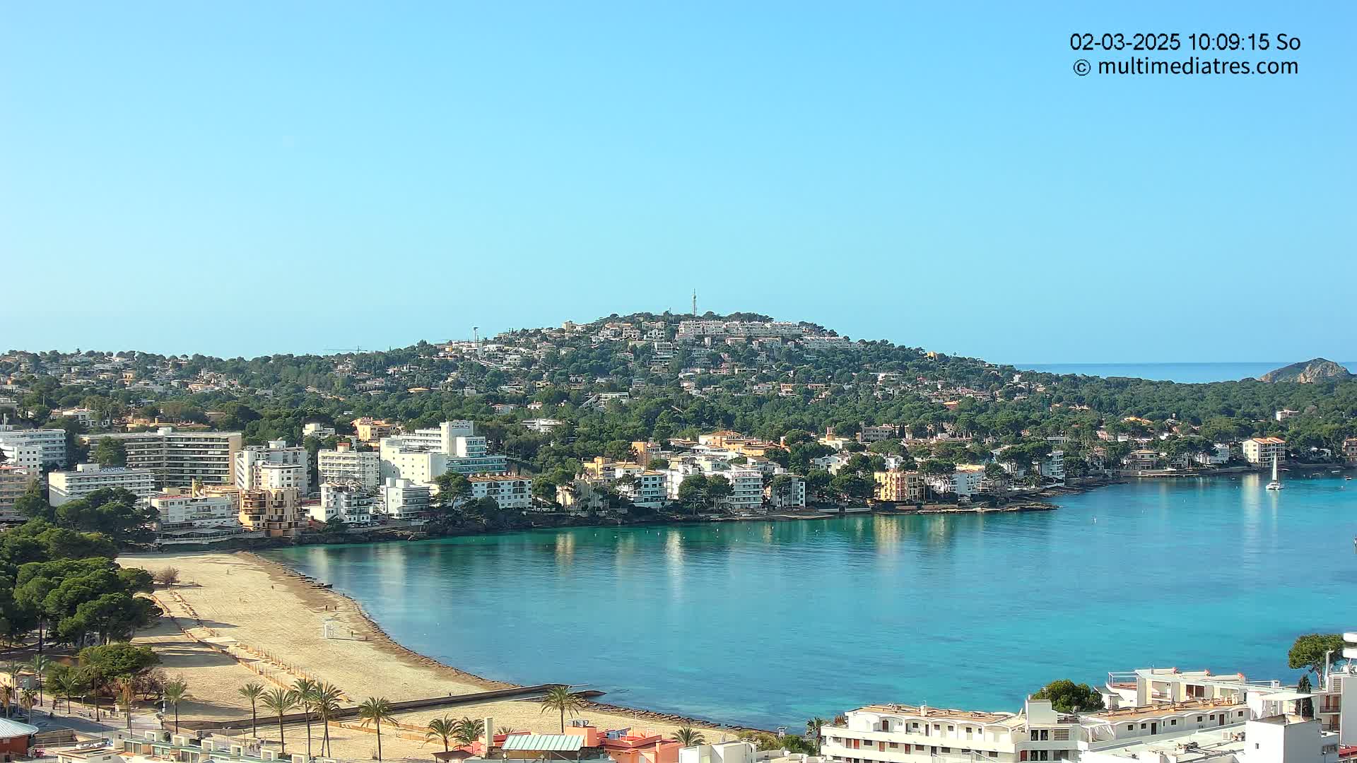 A clear, sunny day overlooks a coastal town nestled against a hillside, featuring a calm, turquoise bay with a sandy beach.