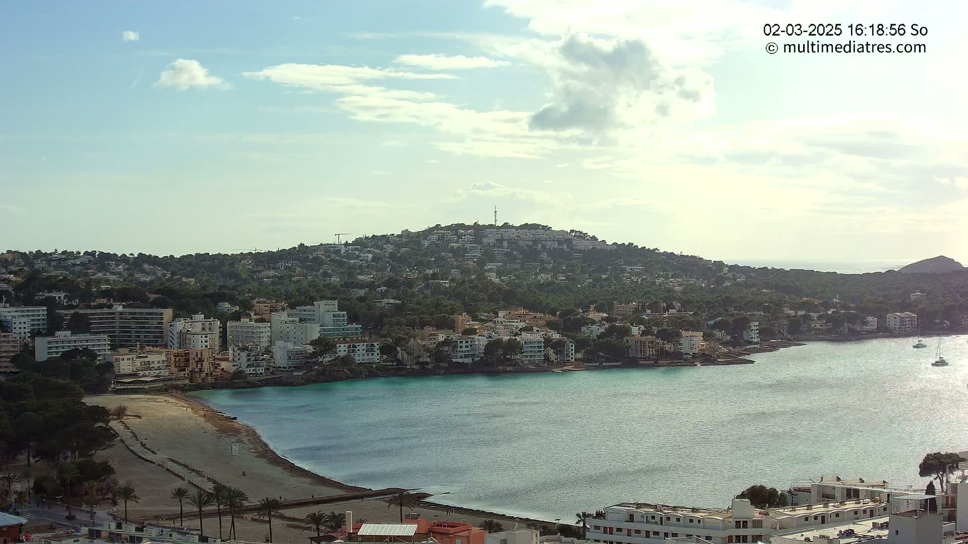A coastal town nestled on a hillside overlooking a calm, turquoise bay under a partly cloudy sky.