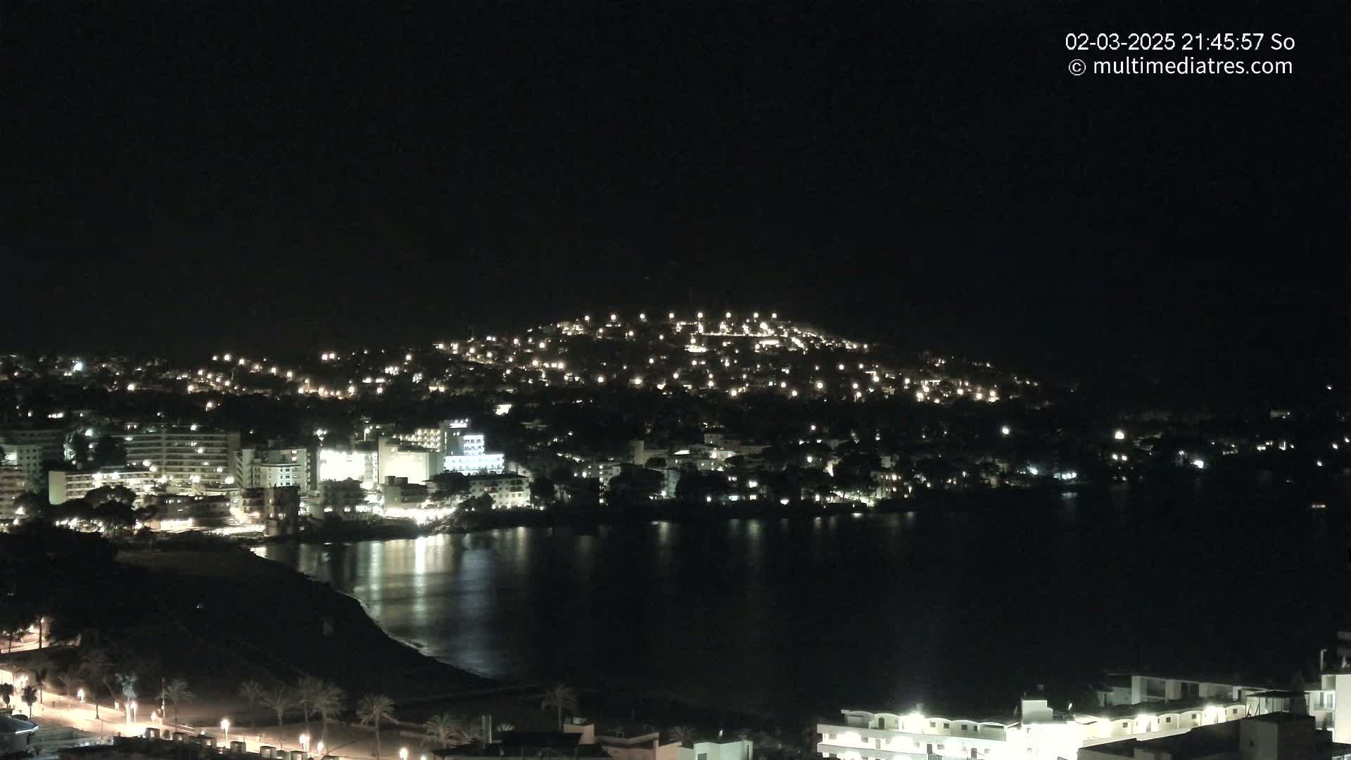 A nighttime view of a coastal town illuminated by lights, nestled against a dark hill, overlooking a calm body of water.
