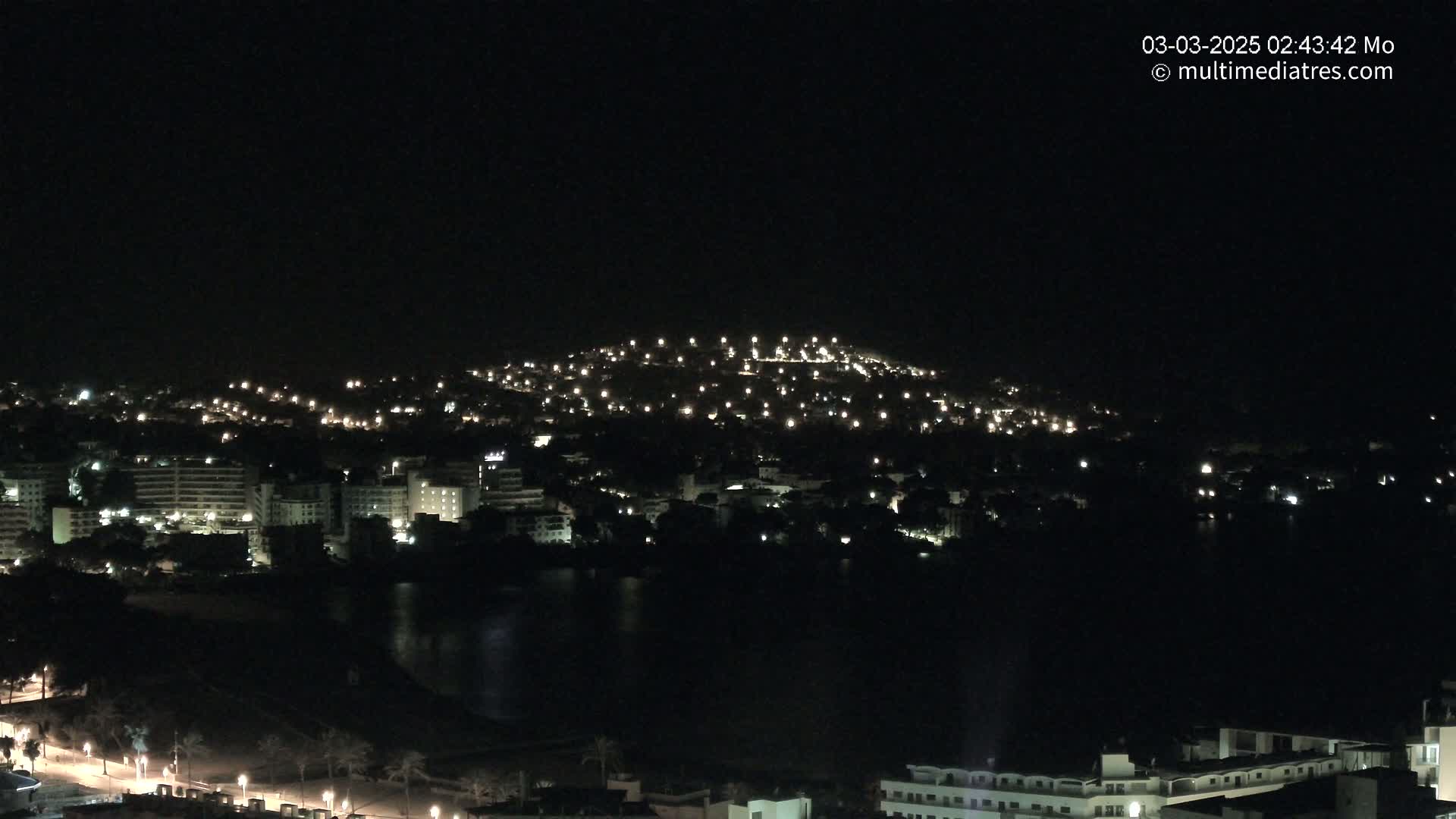 A nighttime view of a city illuminated by scattered lights, nestled beside a dark body of water and a hilltop dotted with lights.