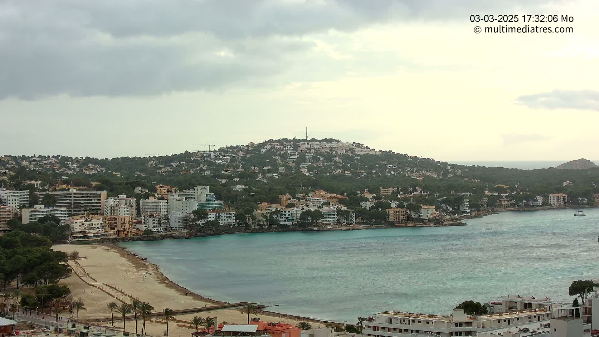 A coastal town with numerous buildings nestled against a hill overlooking a calm, turquoise bay under an overcast sky.