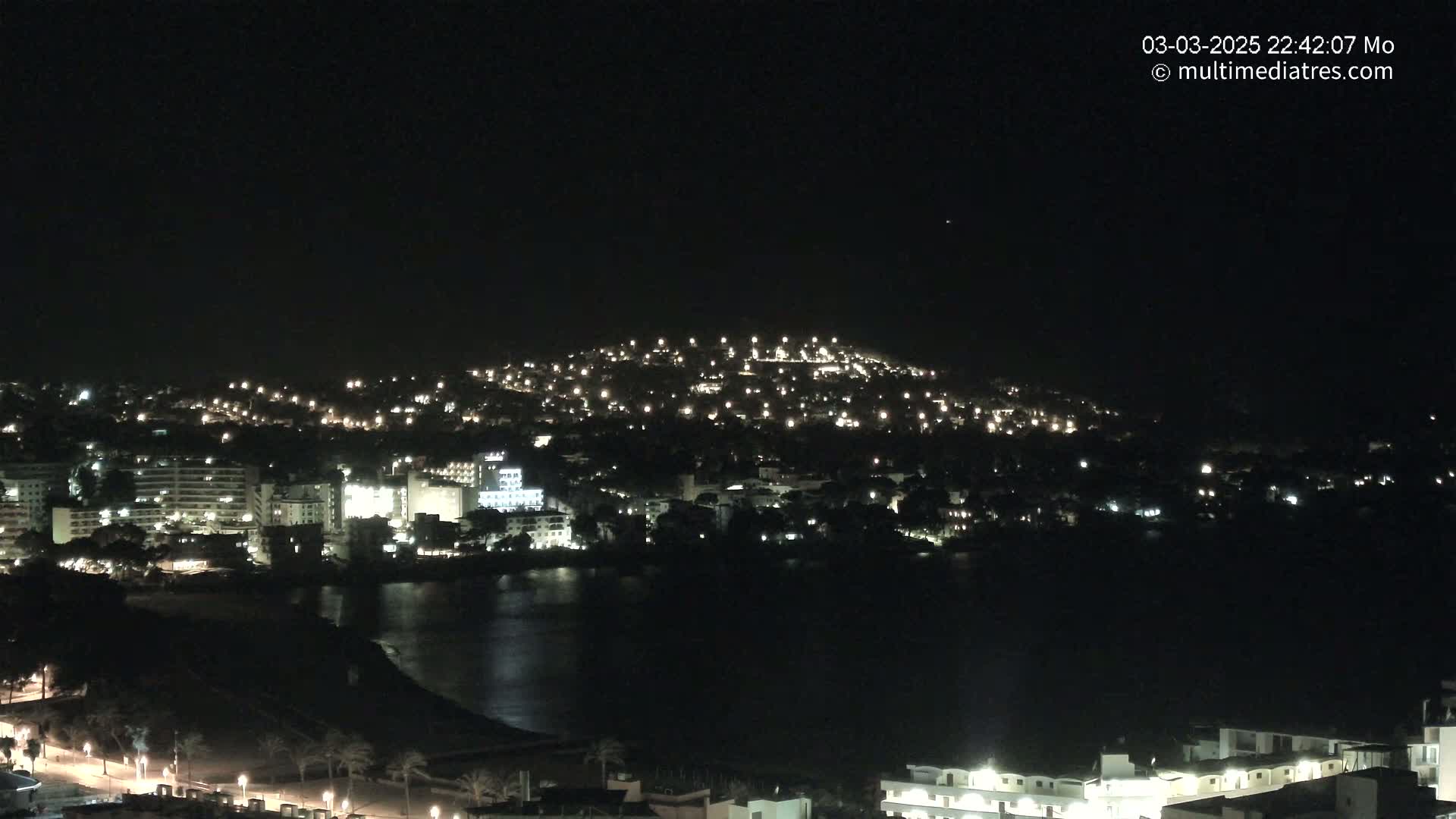 A nighttime view of a coastal town illuminated by lights, with a dark body of water in the foreground under a clear sky.