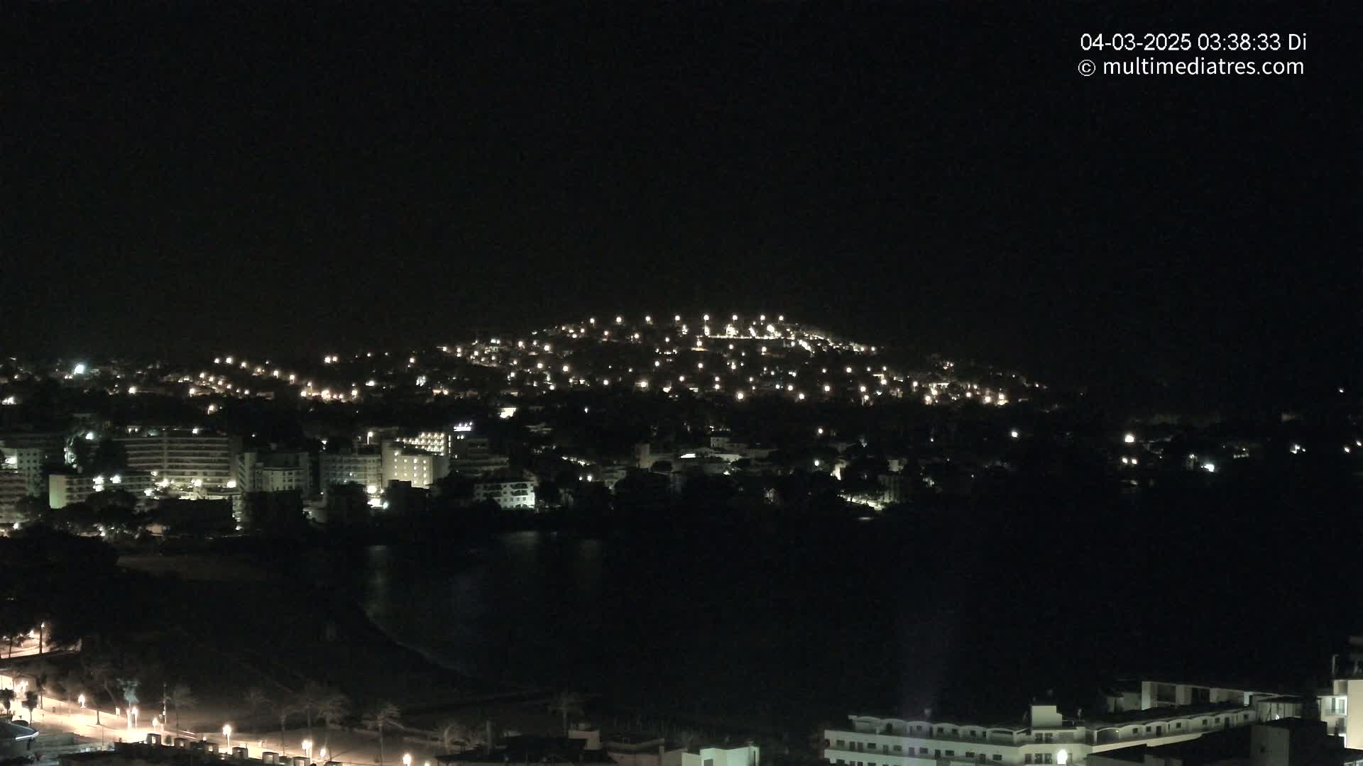 A nighttime view of a city spread across a hill and down to a dark body of water, with numerous lights illuminating buildings and streets.