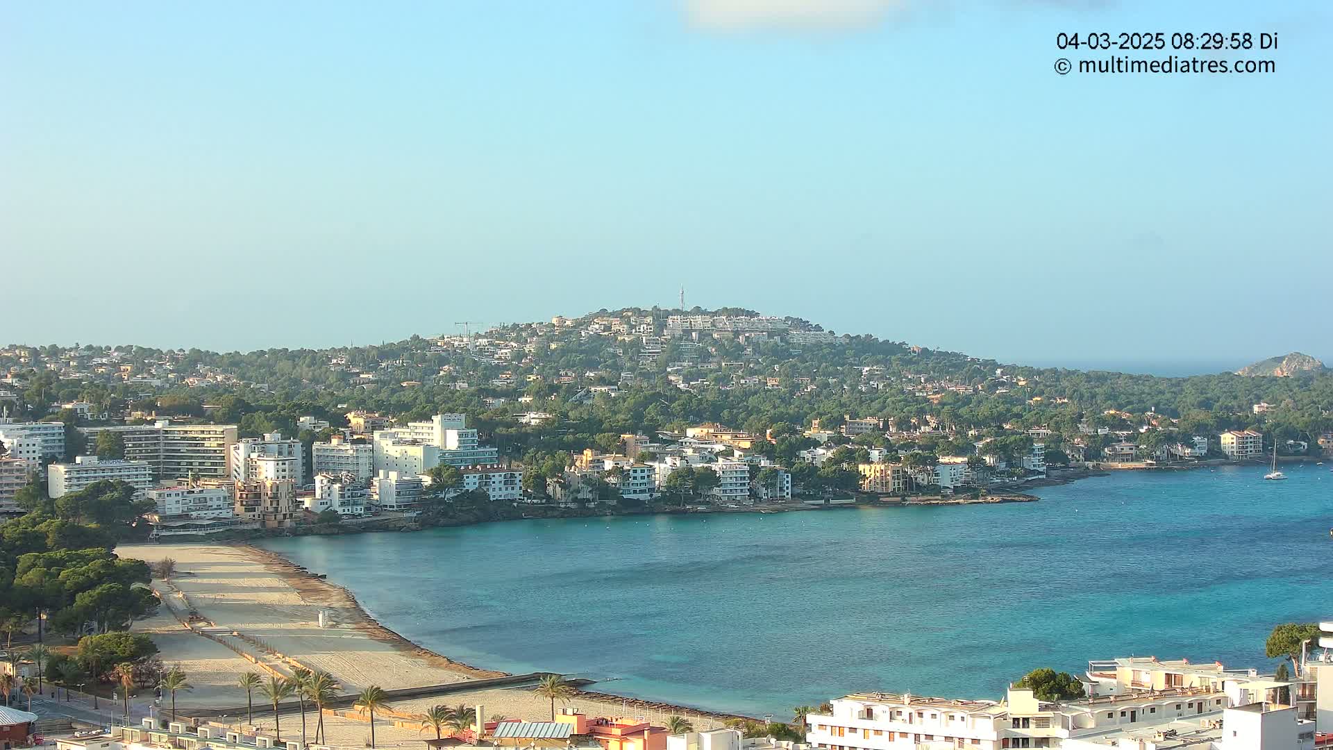 A coastal town on a sunny day, nestled around a calm, turquoise bay with a sandy beach, is backed by a hill covered in lush green vegetation and buildings.