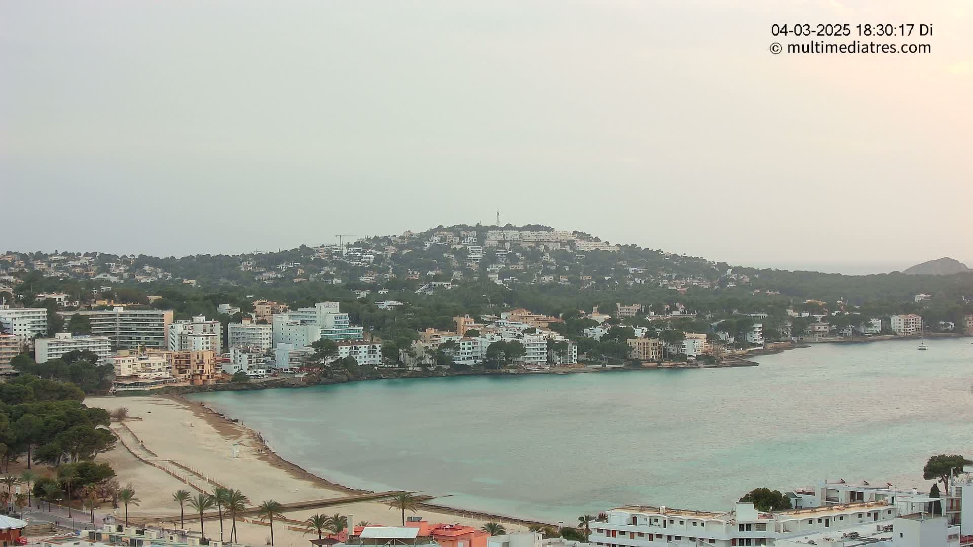 A coastal town on a hill overlooking a calm bay with a sandy beach under an overcast sky.