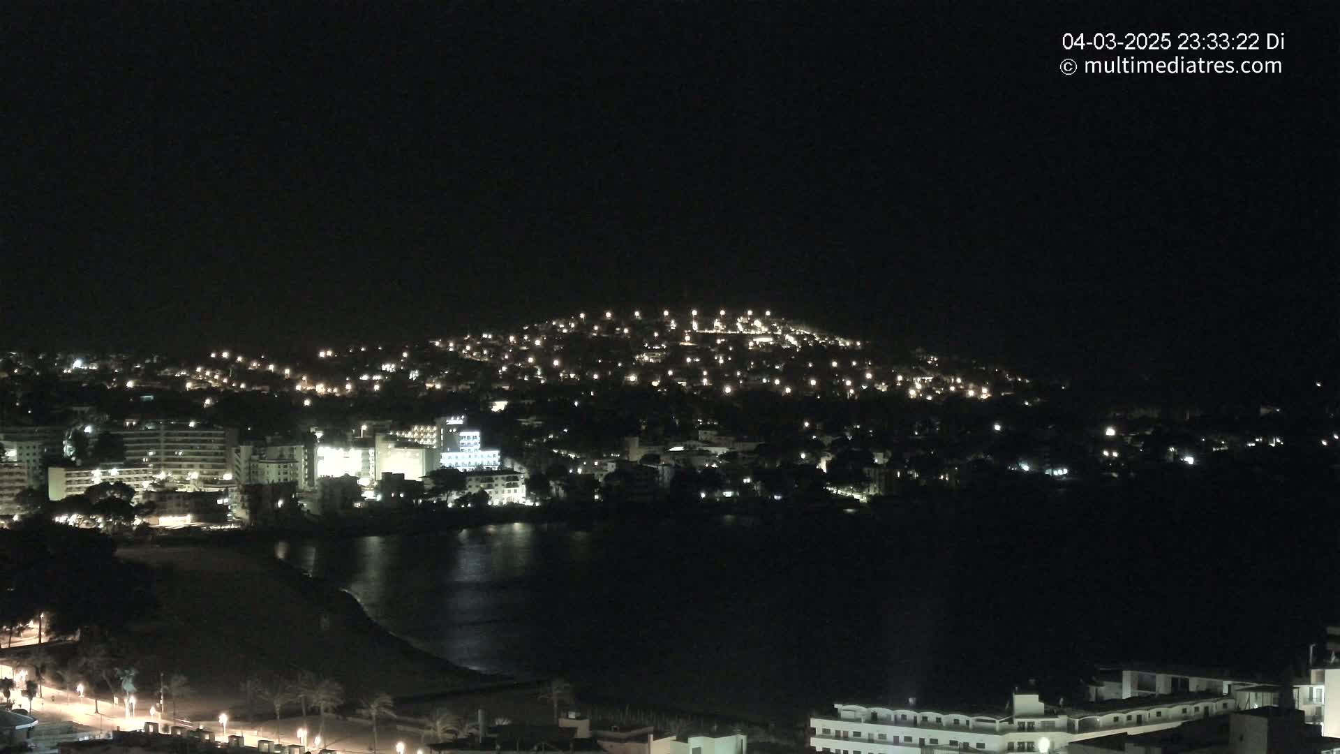 A nighttime coastal town with buildings illuminated along a dark body of water, nestled at the base of a hill also dotted with lights.