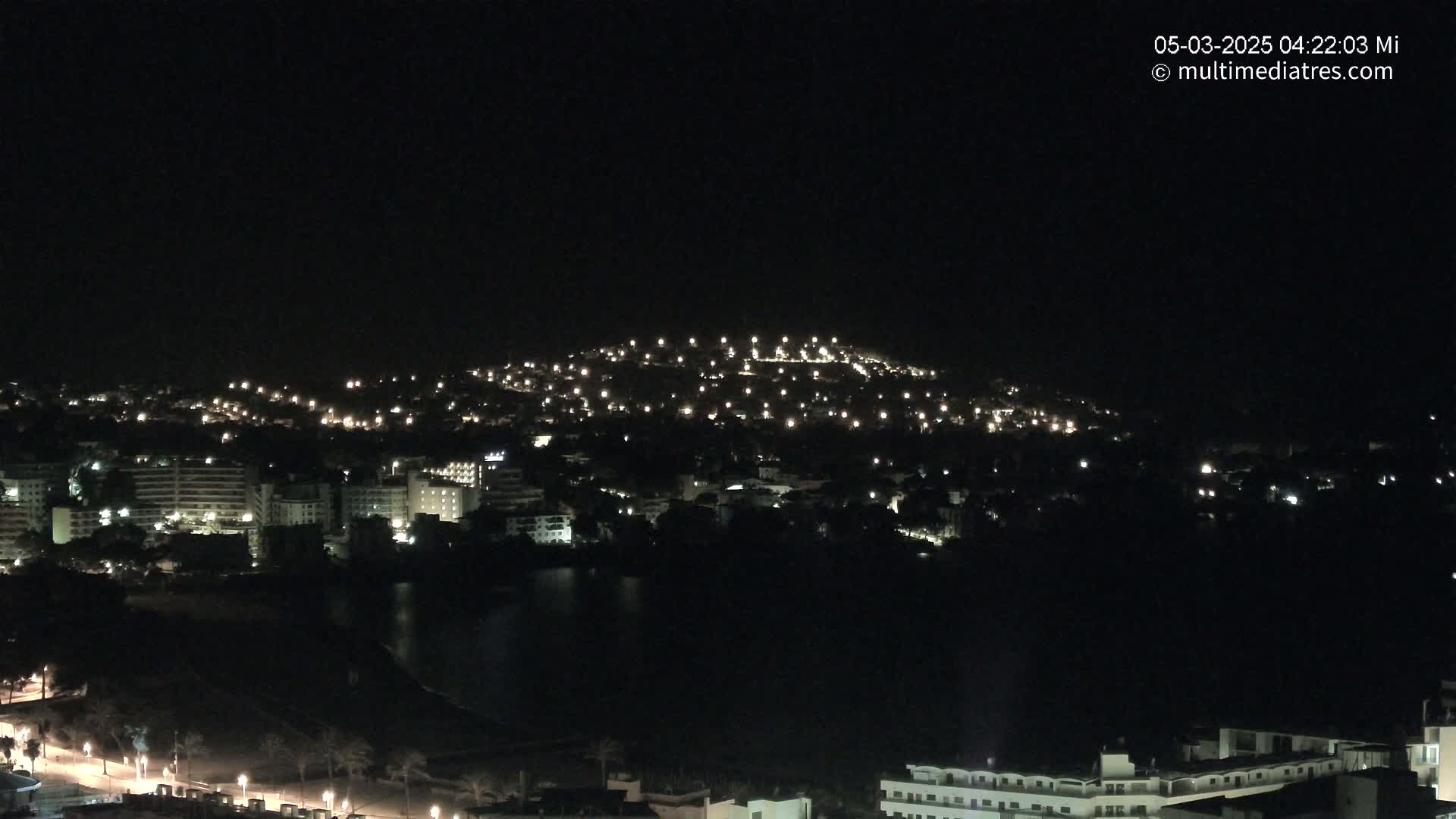 A nighttime view of a city illuminated by numerous lights, situated on a hill next to a dark body of water under a clear, dark sky.
