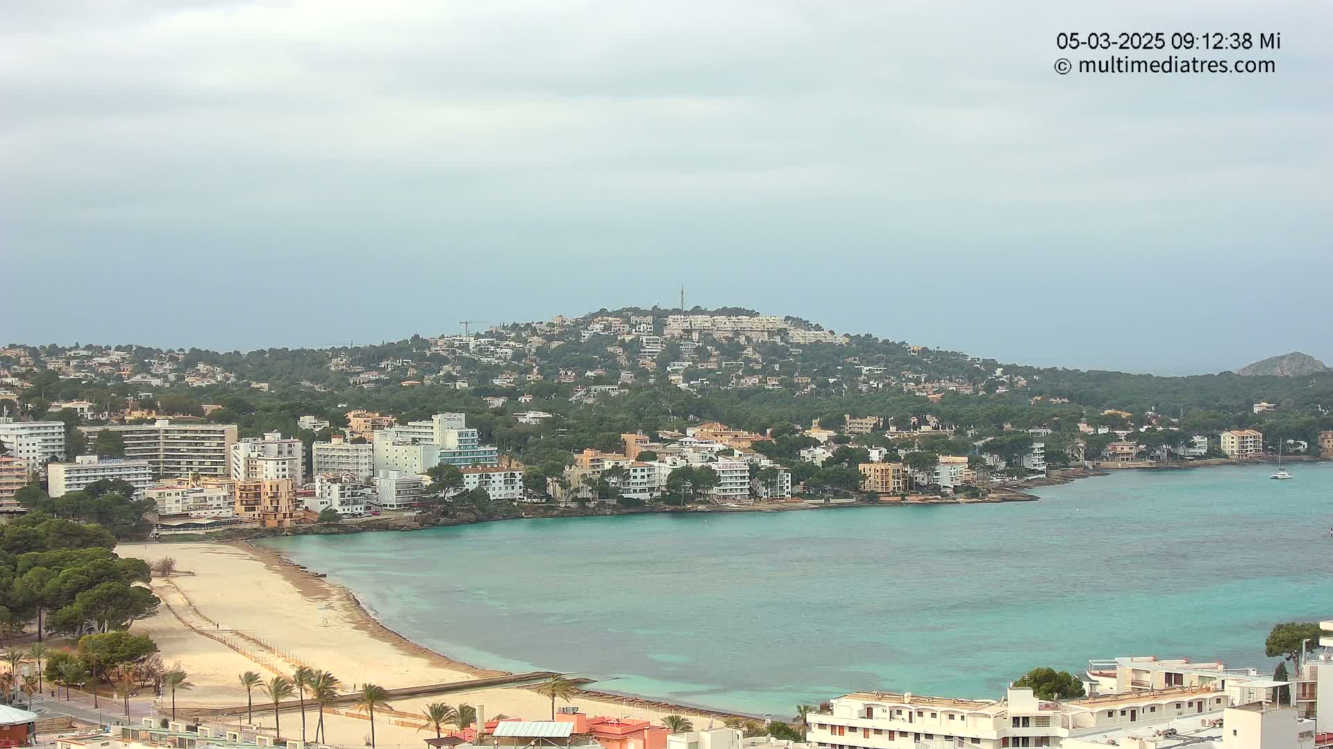 A coastal town with numerous buildings and a sandy beach overlooks a calm, turquoise bay under an overcast sky.