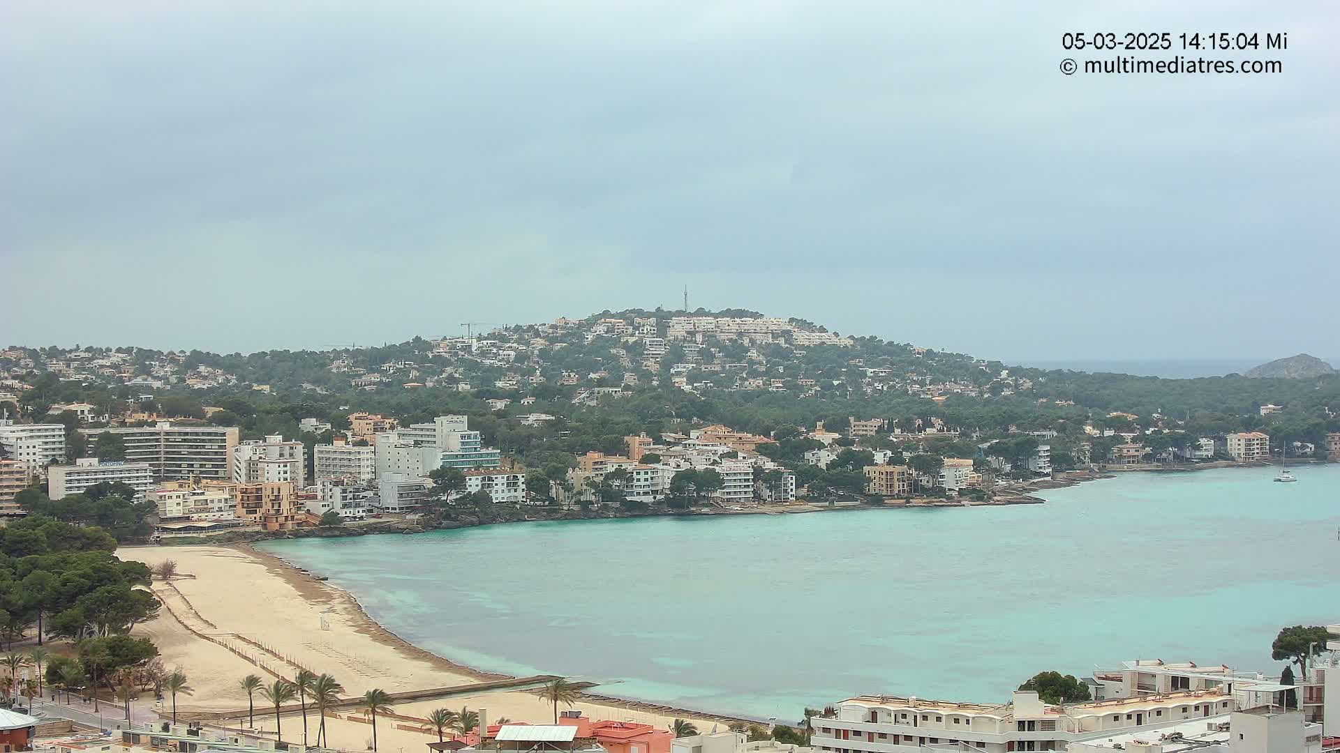 A coastal town with buildings clustered around a sandy beach and turquoise bay, under an overcast sky.
