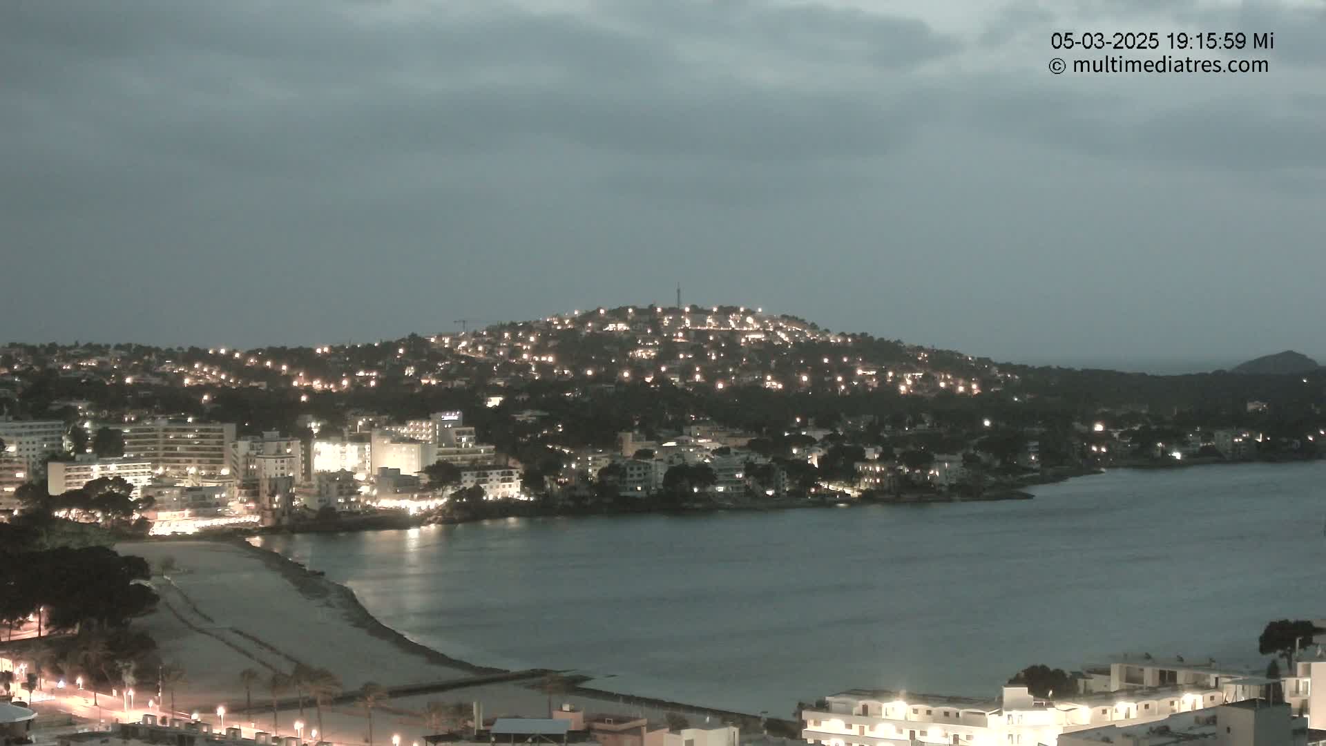 A nighttime coastal town with numerous lights illuminating buildings nestled on a hill overlooking a calm bay and sandy beach under an overcast sky.