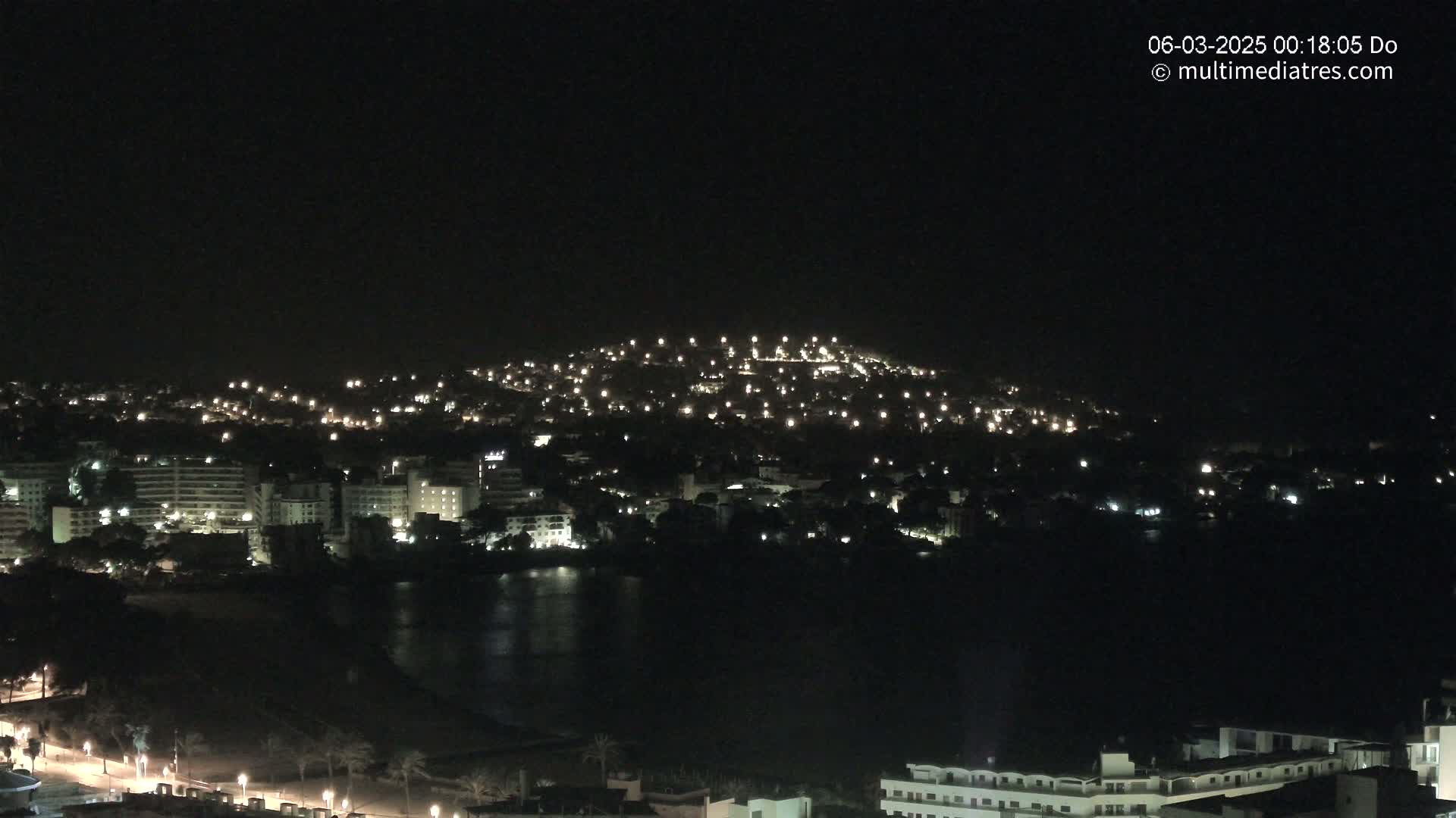 A nighttime view of a coastal town illuminated by many small lights, with a hill in the background and a dark body of water in the foreground.
