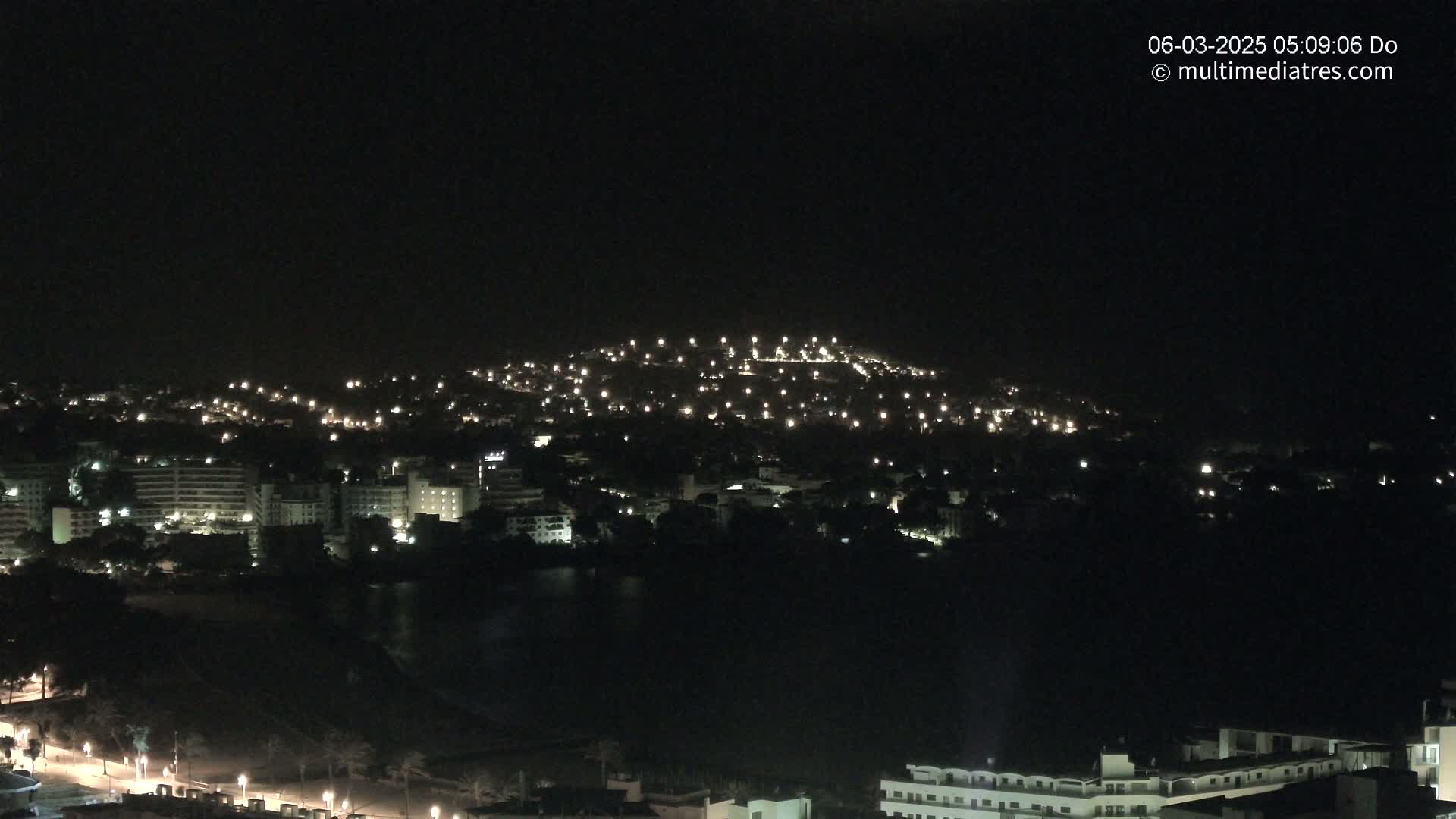 A nighttime view of a coastal town illuminated by many scattered lights, situated on a hill overlooking a dark body of water.