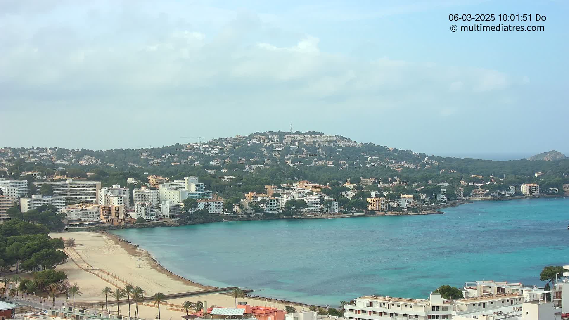 A coastal town with a sandy beach and turquoise water sits beneath a hill covered in buildings and trees under a partly cloudy sky.