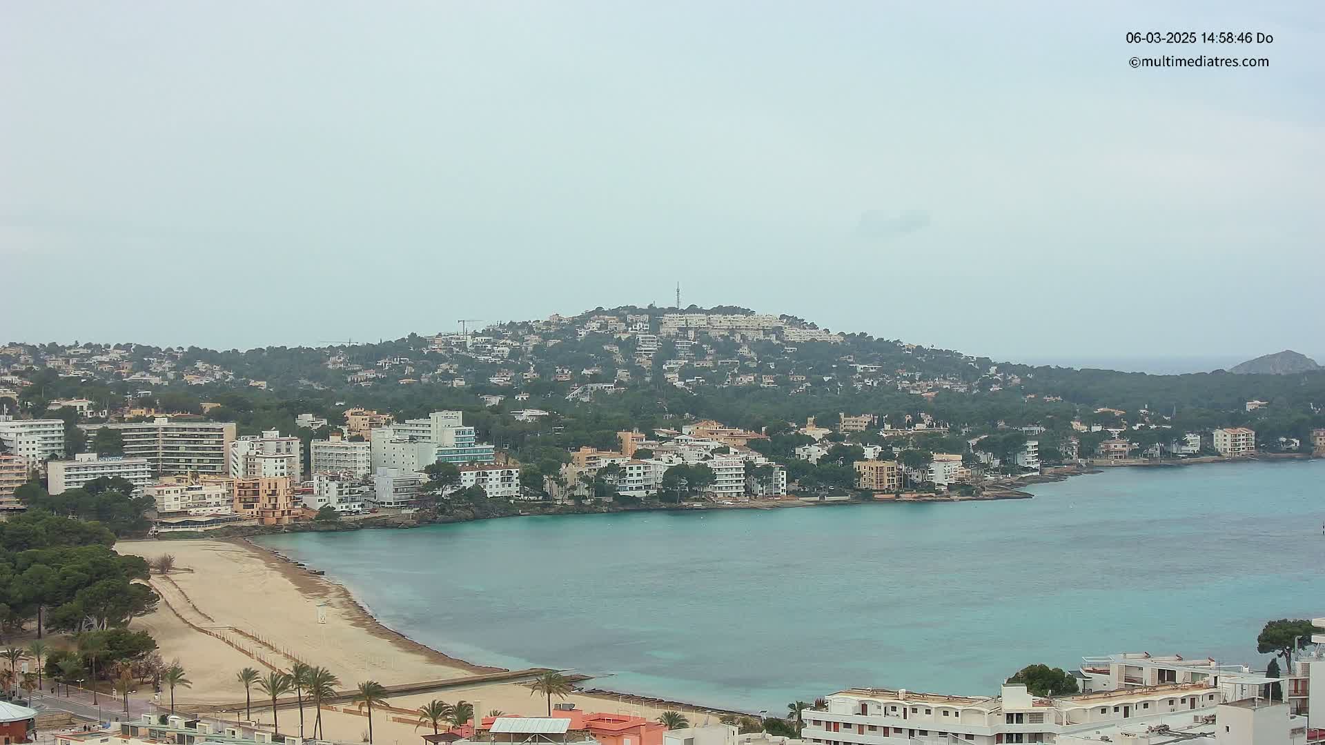 A coastal town with numerous buildings, a sandy beach, and turquoise water under an overcast sky.
