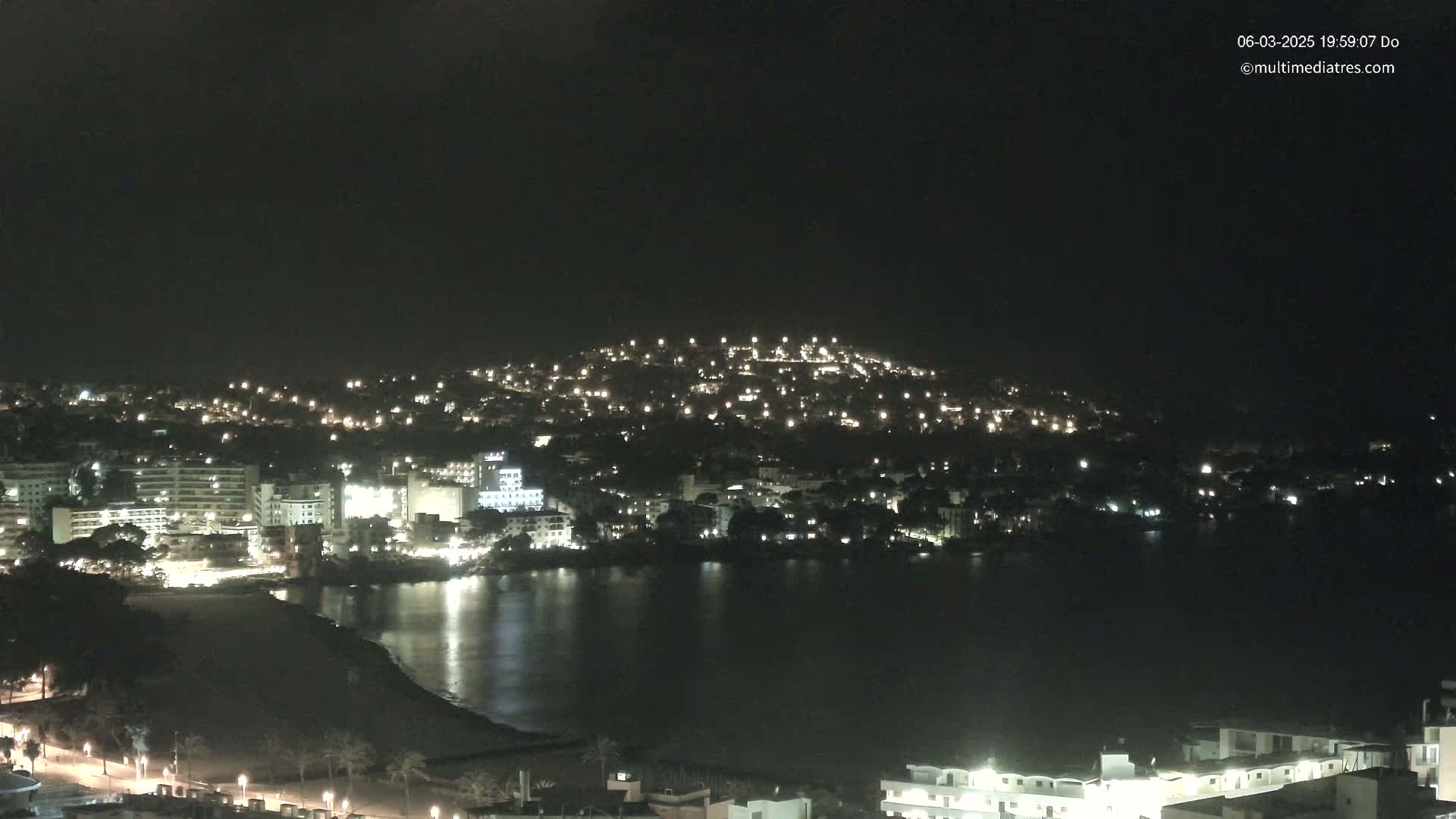 A nighttime view of a coastal town with buildings illuminated along a calm body of water, under a clear, dark sky.