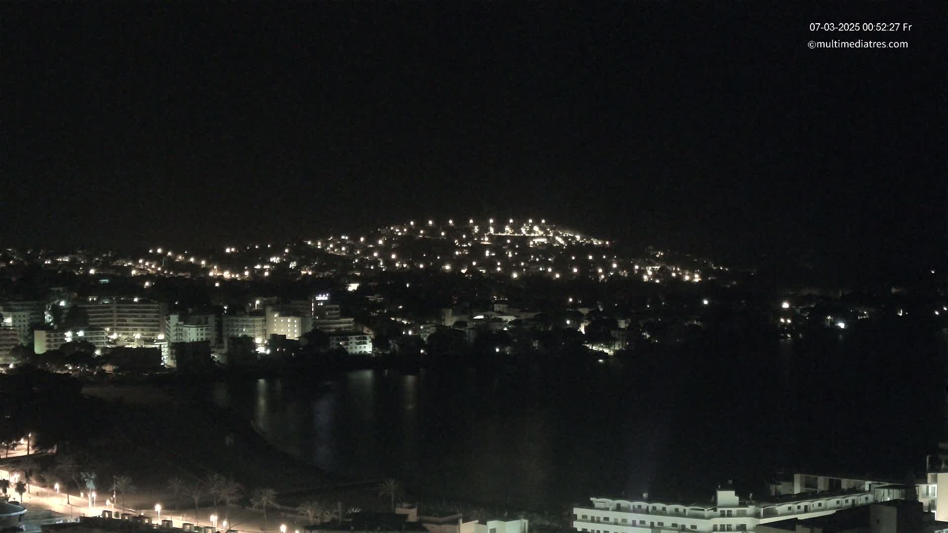 A nighttime view of a city illuminated by lights, nestled beside a dark body of water, with a hilltop also speckled with lights in the background under a clear, dark sky.
